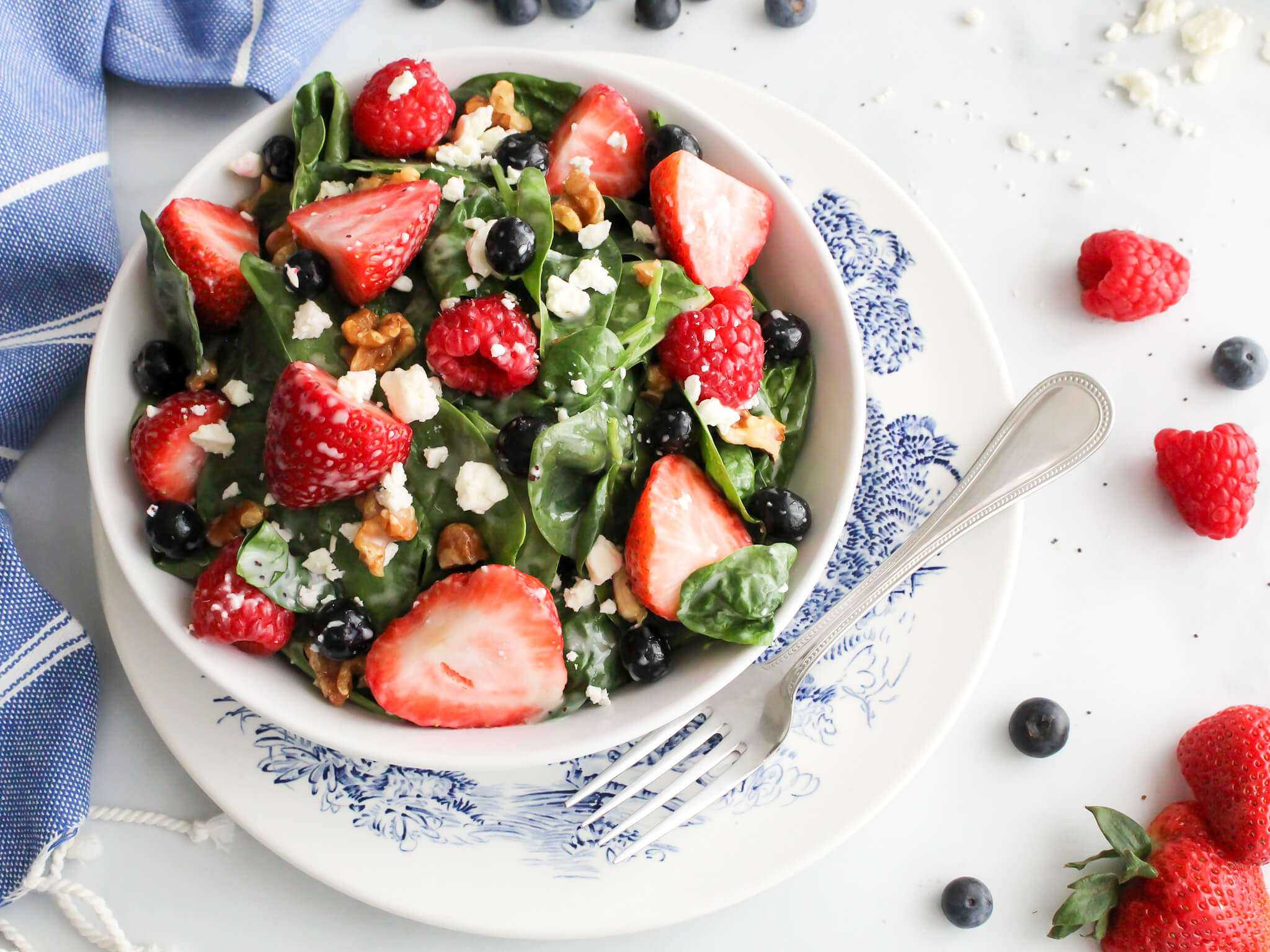 Bowl of fresh spinach strawberry and bluberry salad with poppy seed dressing topped with feta cheese and walnuts served on top of a blue and white transferware plate