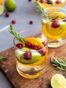 Cocktail glasses filled with sparkling white sangria with cranberries, orange and lime slices, bits of apple, and garnished with a sprig of fresh rosemary