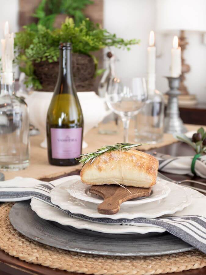 Dining room table set for an Italian-themed dinner party with a rustic bread board on a plate and a piece of ciabatta bread wrapped in twine and fresh herbs