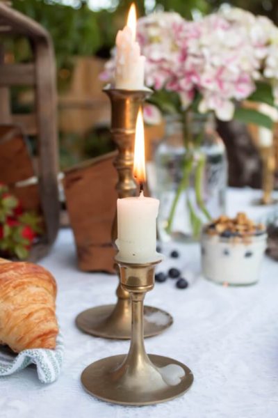 Close up of vintage brass candlesticks on the table setting for a backyard summer picnic with flowers and breakfast food in the background