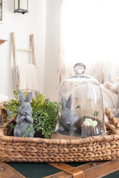 Cozy living room Easter vignette featuring a woven tray, bunny decor, greenery, vintage letters, and speckled eggs under a glass cloche.