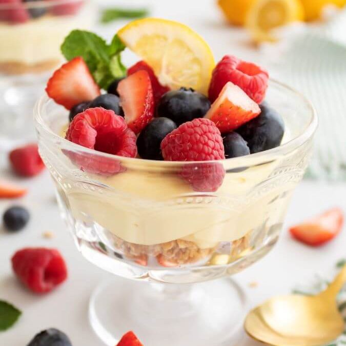 No-bake lemon cheesecake cups tipped with fresh raspberries, blueberries, and strawberries slices and garnished with a slice of lemon and sprig of fresh mint