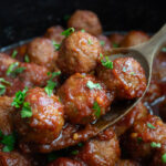 Close up view of Crockpot Bourbon BBQ Meatballs in the slow cooker displayed on a wooden spoon