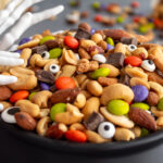 Close up of Halloween Monster Munch Trail Mix with candy eye sprinkles in a caldron bowl with spooky skeleton hand serving utensils on the side of the caldron