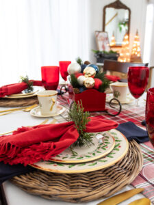 Red and blue Christmas tablescape set for the holidays with Lenox Christmas china, layered place settings, red napkins, gold silverware, red wine goblets, and a Santa' sleigh centerpiece filled with Christmas ornaments and greenery