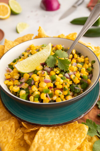 Bowl of chili roasted corn salsa garnished with a lime slice and fresh cilantro displayed on top of a turqouise colored plate surrounded by yellow tortilla chips