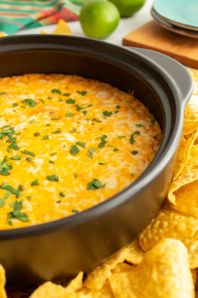 Side view of cream cheese bean dip in a black crock with melted Mexican cheese on top, a cilantro garnish and surrounded by yellow tortilla chips