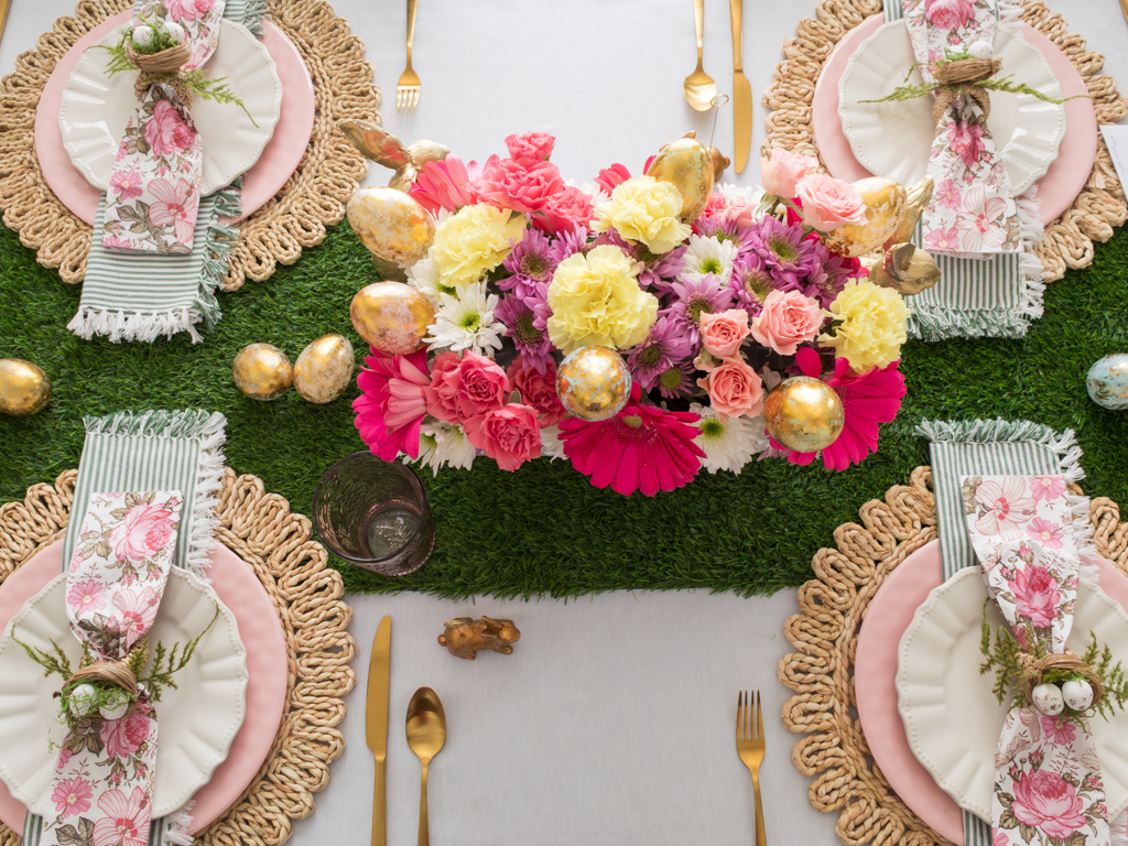 Overhead view of a colorful DIY Easter floral centerpiece with faux eggs set on a dining room table styled for Easter brunch with a faux grass table runner, rattan charger plates, colorful plates and linens and gold silverware. 