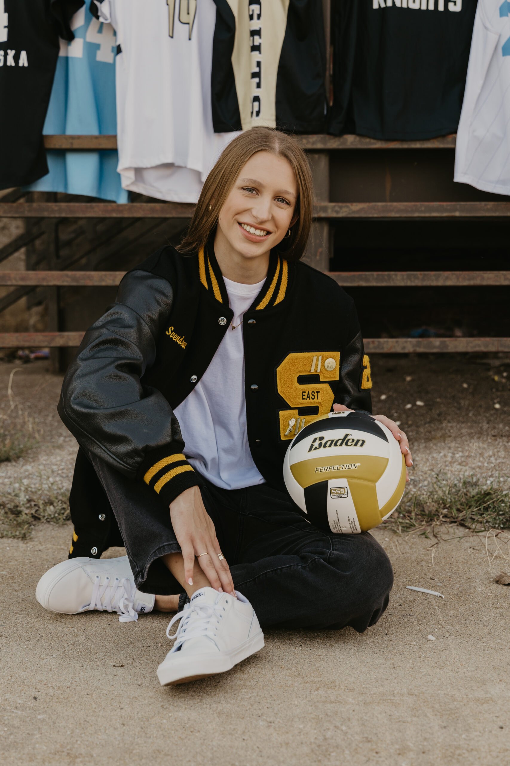 Girl in letter jacket holding a volleyball for senior photos with other sports jerseys hanging in the background