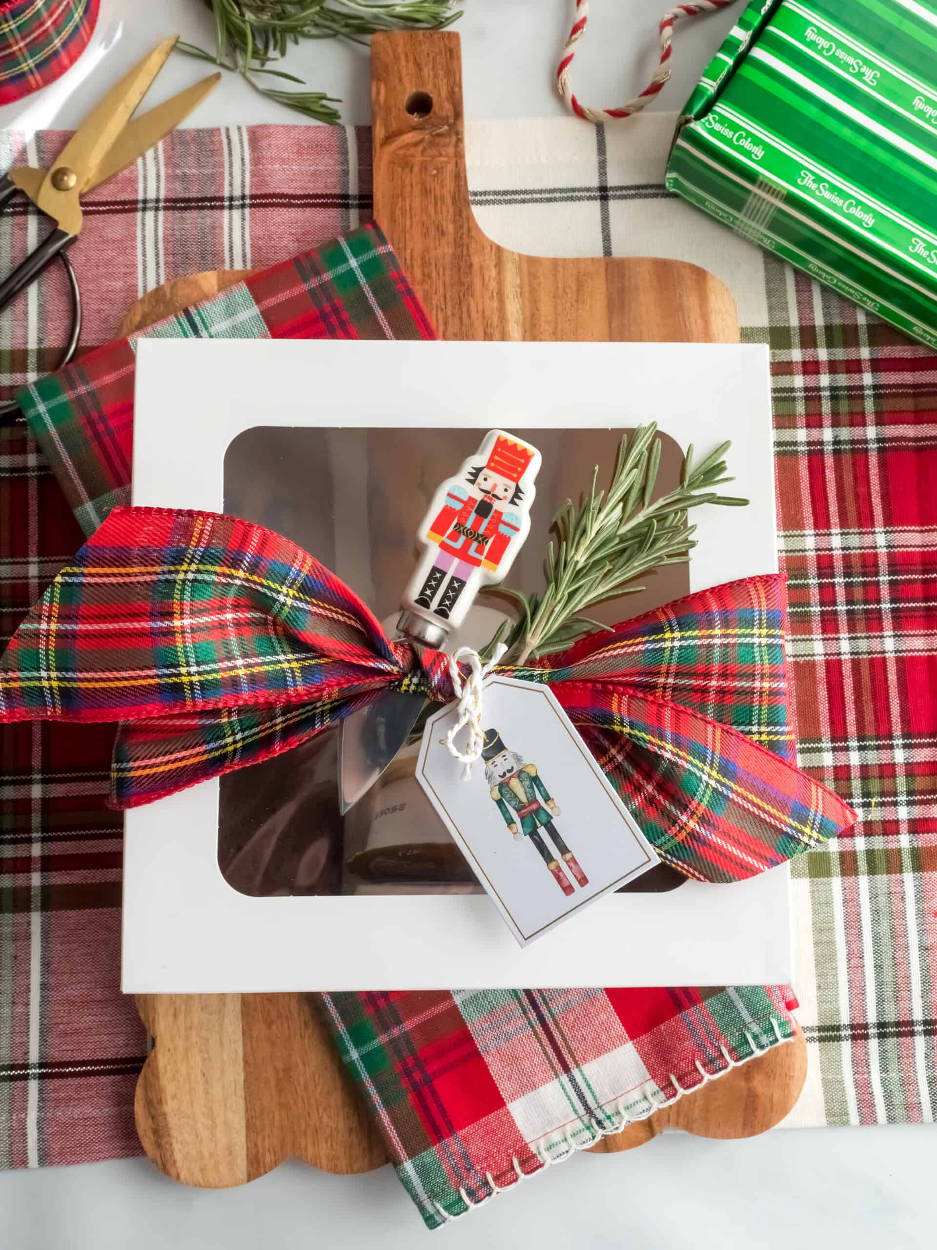 A white bakery box wrapped with a plaid bow and a spring of fresh rosemary with a nutcrack gift tag and a nutcracker cheese knife tucked into the ribbon. The box is is packaged as a christas gift with a plaid tea towel and a scalloped wooden cutting board and the box is filled with mini breads ands sweet cremes from Swiss Colony. 