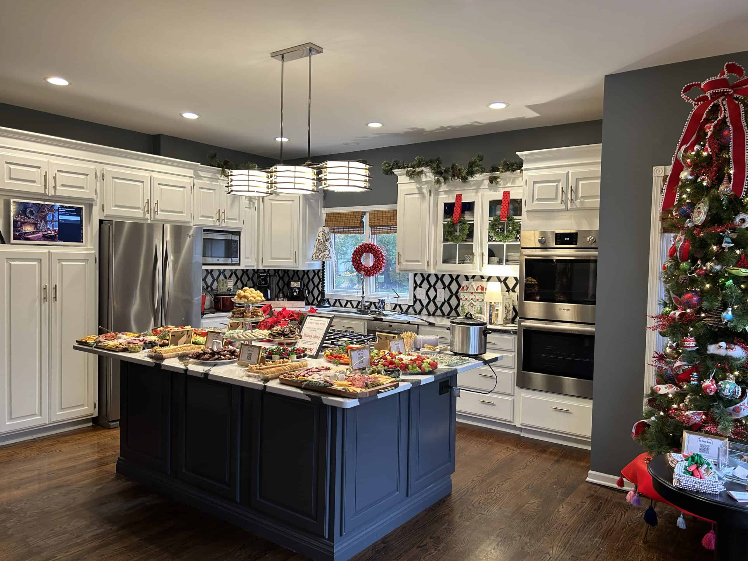 Kitchen decorated for Christmas with a tree in the corner and wreaths on the cabinets and a spread of holiday party food on the island. 