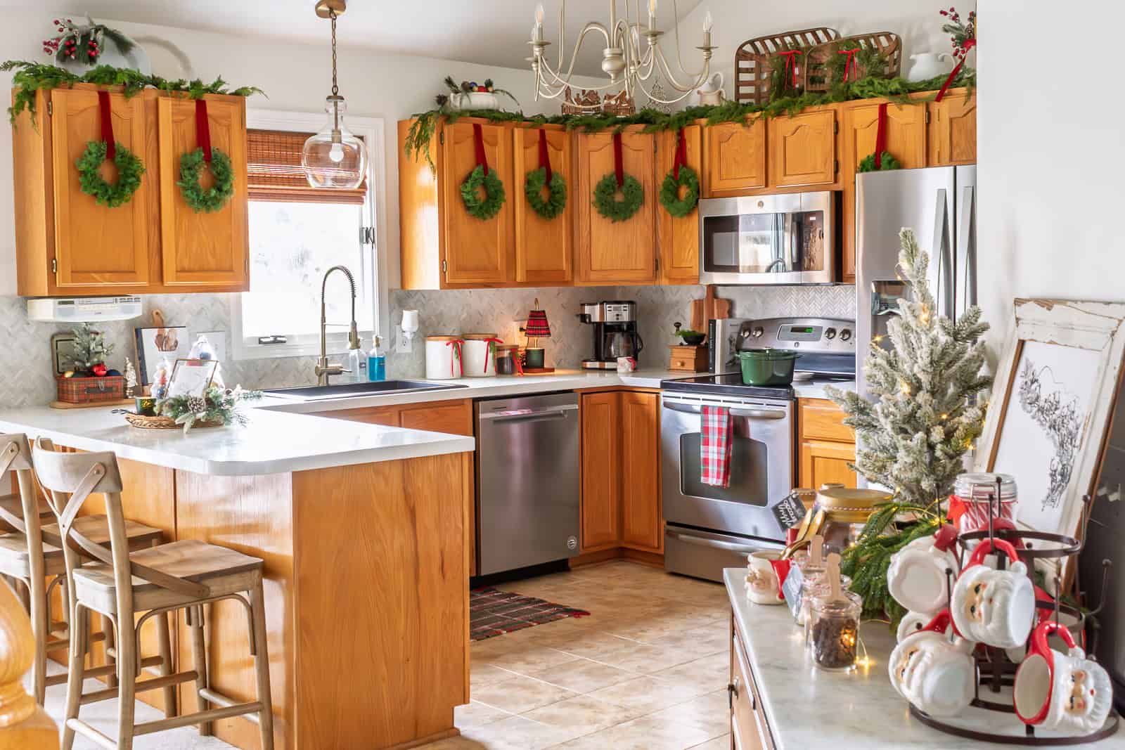 Kitchen decorated with traditional Christmas decorating ideas, featuring wreaths on cabinet doors with red velvet ribbon, garland above the cabinets, tartan accents, vintage cottage-style vignettes, and warm metals for a Ralph Lauren–inspired Christmas look.