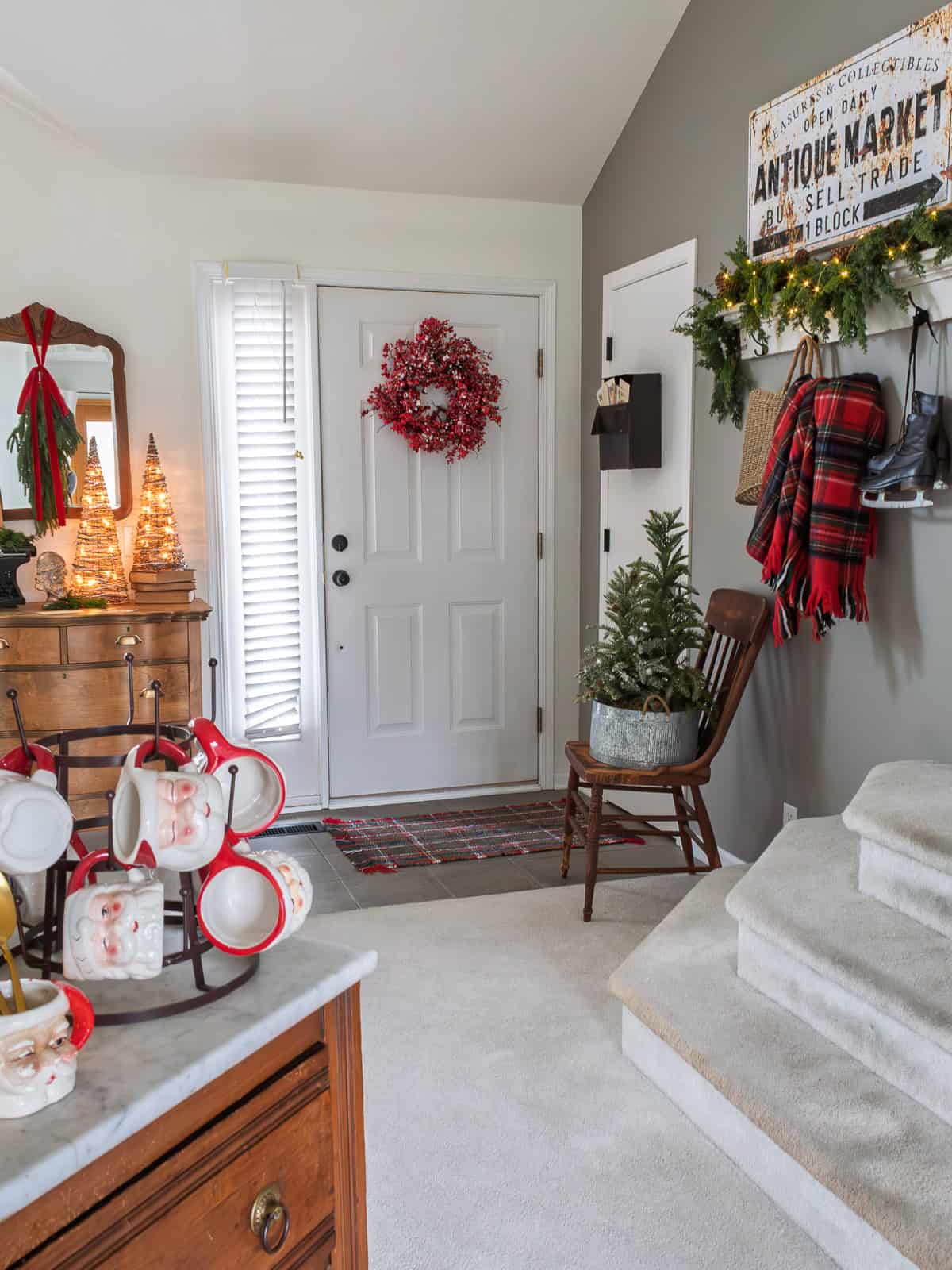 Traditional Christmas entryway decorated with a flocked mini tree on a vintage wooden chair, a red berry wreath on the front door, plaid scarf and boots on wall hooks, vintage dresser with Santa mugs on display, and warm holiday greenery creating a cozy and welcoming Christmas foyer.