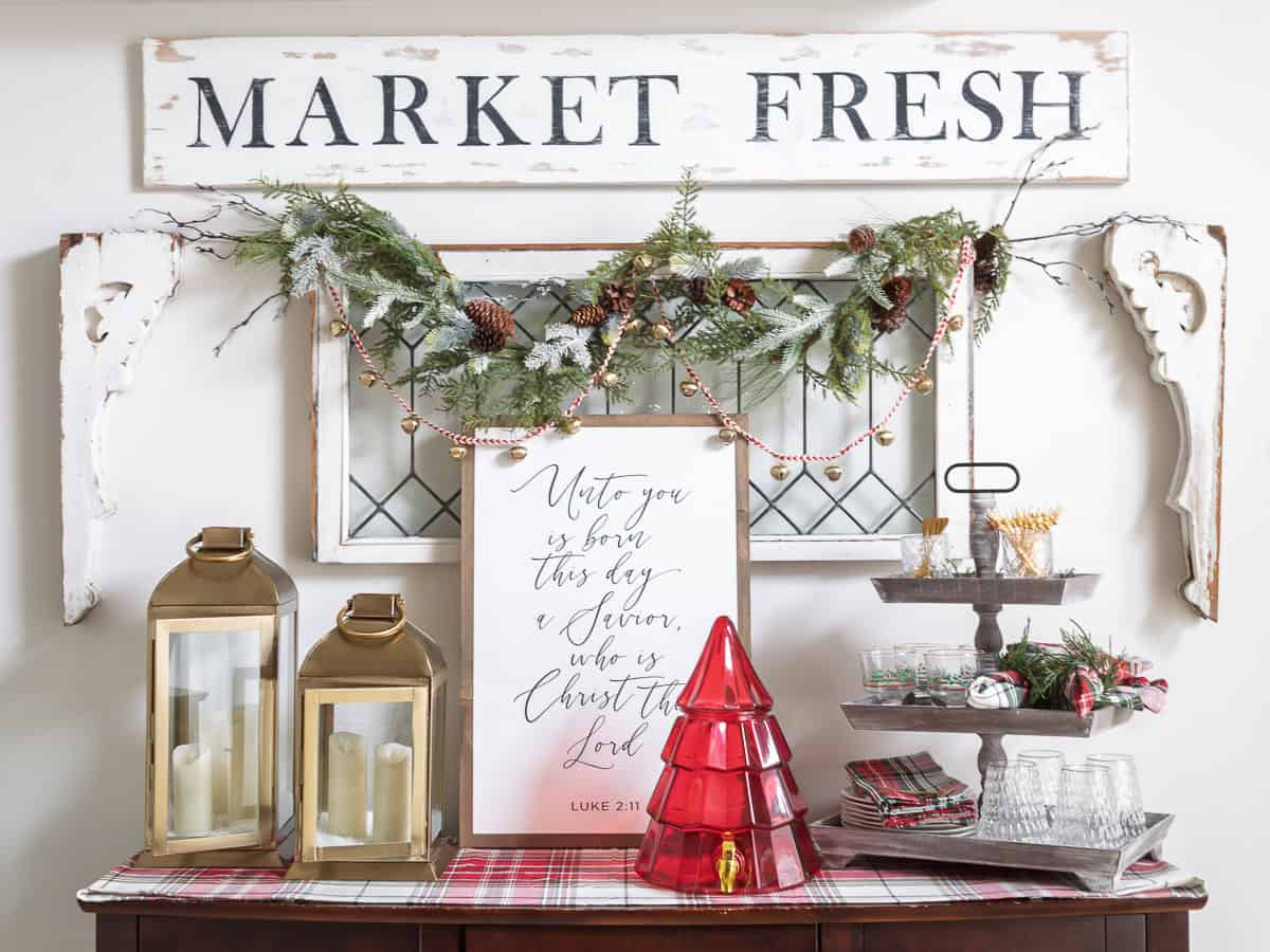 Christmas buffet decorated with traditional holiday accents, including a red tree-shaped drink dispenser, gold lanterns with candles, a Luke 2:11 Christmas sign, layered plaid runner, vintage corbels, farmhouse window frame, and greenery garland with bells, creating a classic and traditional Christmas decorating look in the dining room.