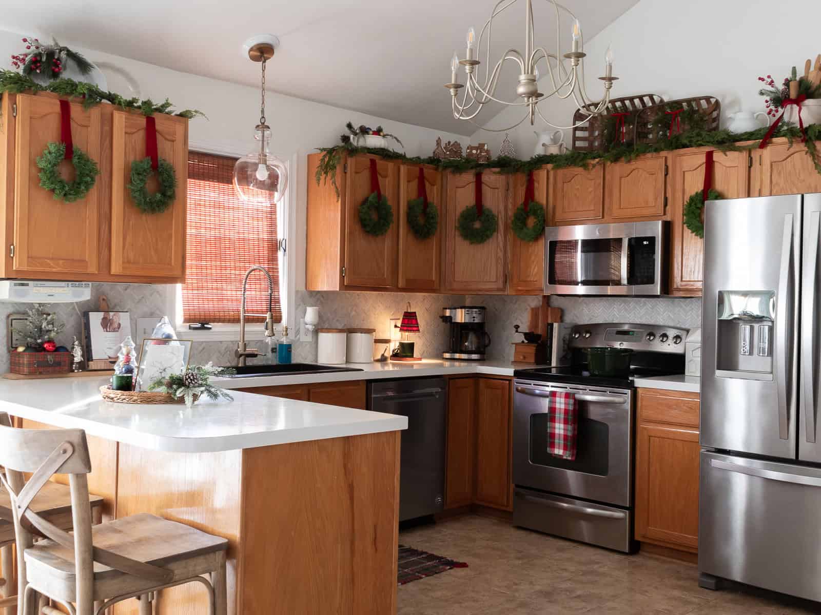 “Full kitchen decorated with traditional Christmas decorating ideas, including wreaths hung on cabinet doors with red velvet ribbon, garland and gingerbread village houses above the cupboards, warm wood cabinets, plaid accents, a green Dutch oven on the stove, and classic Ralph Lauren Christmas inspired touches throughout the space.