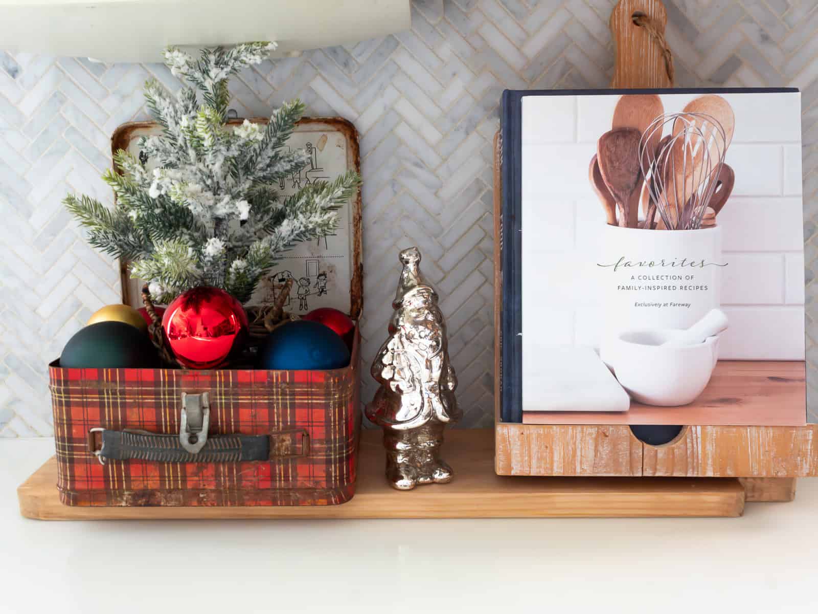 vintage plaid lunch box filled with ornaments and a flocked mini Christmas tree styled on a cutting board beside a silver tree figurine and cookbook holder, creating a cozy vignette that fits traditional Christmas decorating ideas and a classic Ralph Lauren Christmas look