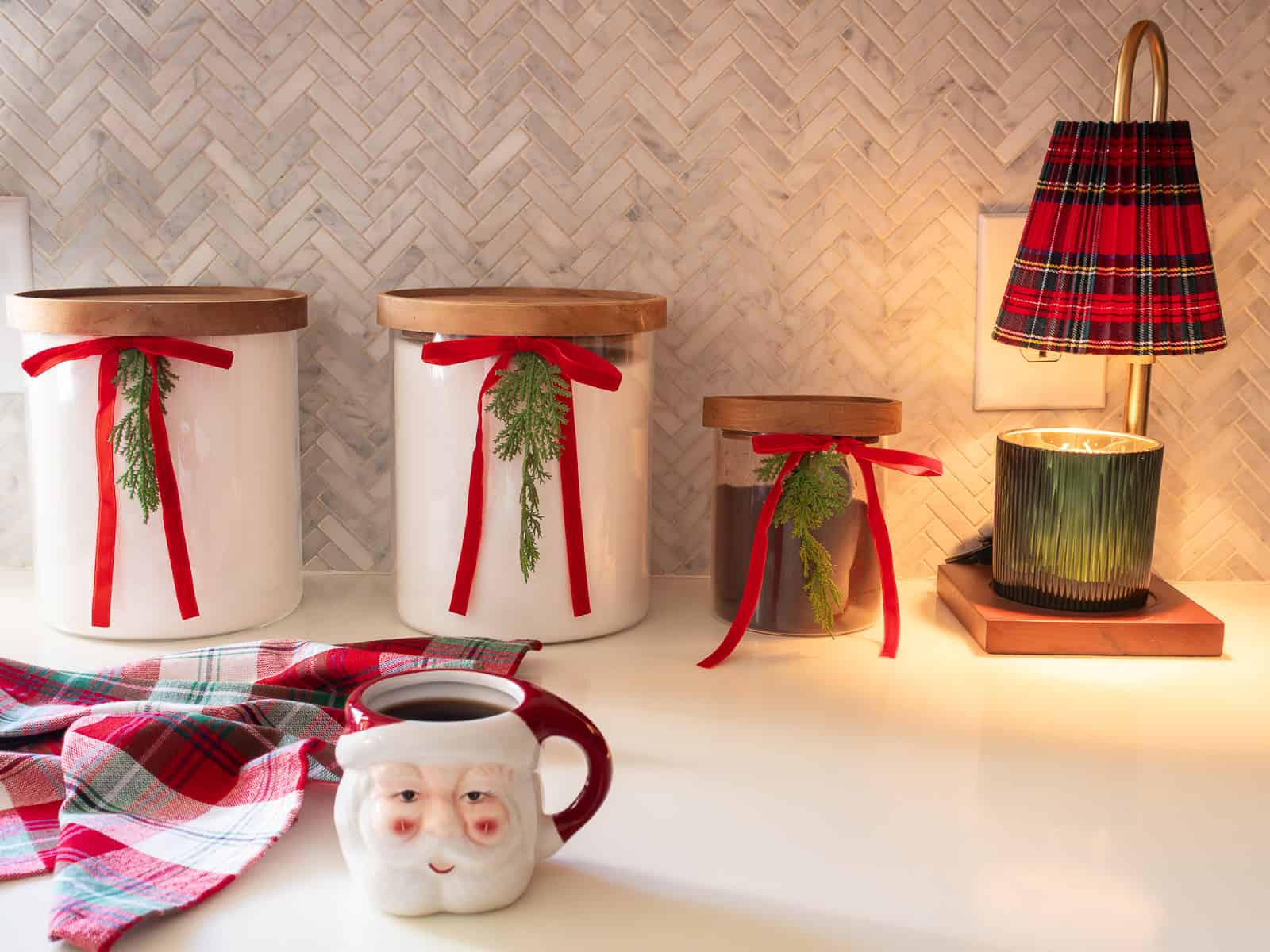 glass kichen cannisters with wooden lids filled with sugar, coffee, and tea, decorated for Christmas with a spring of greenery and red velevet ribbon. A santa mug in the foreground filled with coffee next to a plaid Christmas tea towle and a plaid candle warmer nearby