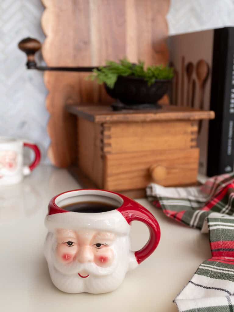 Santa mug filled with coffee styled on a kitchen counter with plaid napkins, a vintage wooden coffee grinder, greenery, and cutting boards, creating a cozy vignette that fits traditional Christmas decorating ideas and classic Ralph Lauren Christmas style.