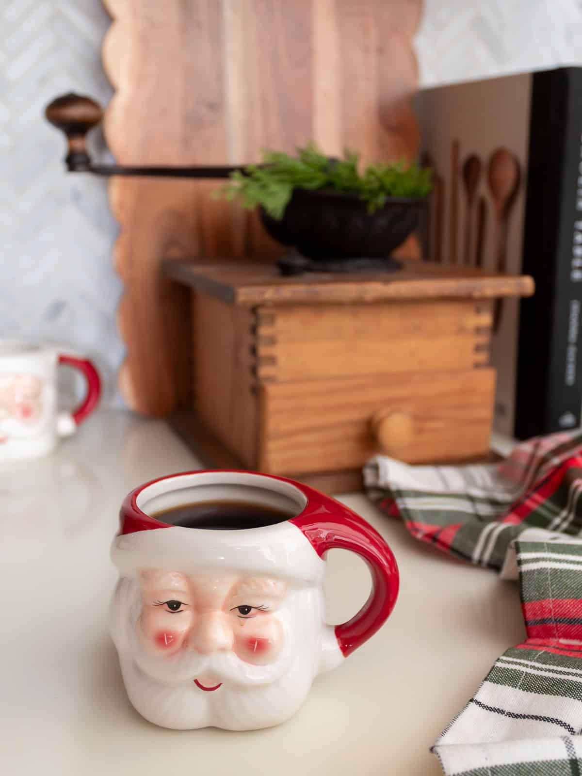 Santa mug filled with coffee styled on a kitchen counter with plaid napkins, a vintage wooden coffee grinder, greenery, and cutting boards, creating a cozy vignette that fits traditional Christmas decorating ideas and classic Ralph Lauren Christmas style.