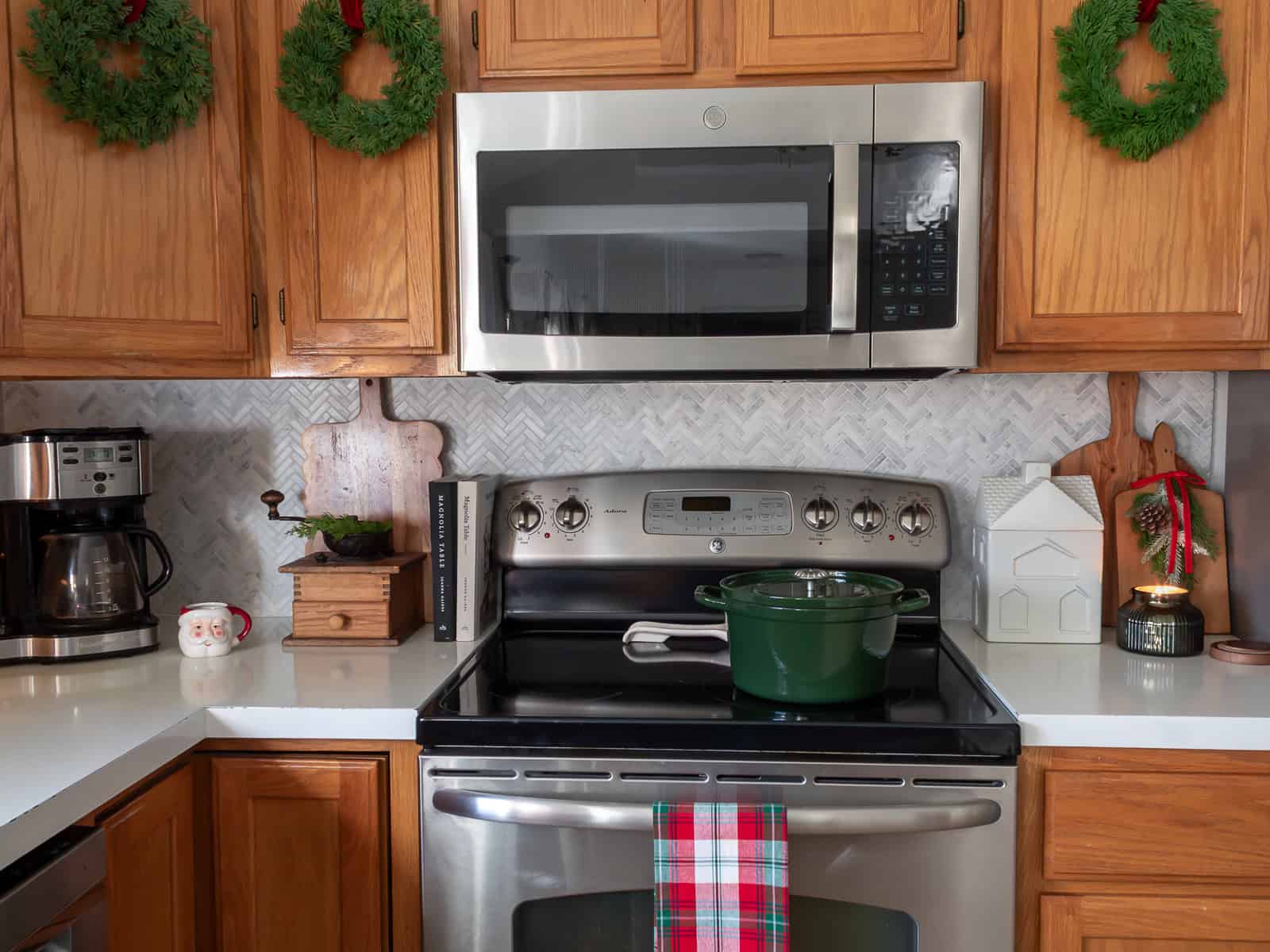 Stove area decorated with traditional Christmas decorating ideas, including wreaths hung on cabinet doors with red velvet ribbon, layered cutting boards, a vintage coffee grinder with greenery, a white ceramic house, a green Dutch oven, and plaid towel for a cozy Ralph Lauren Christmas inspired kitchen.