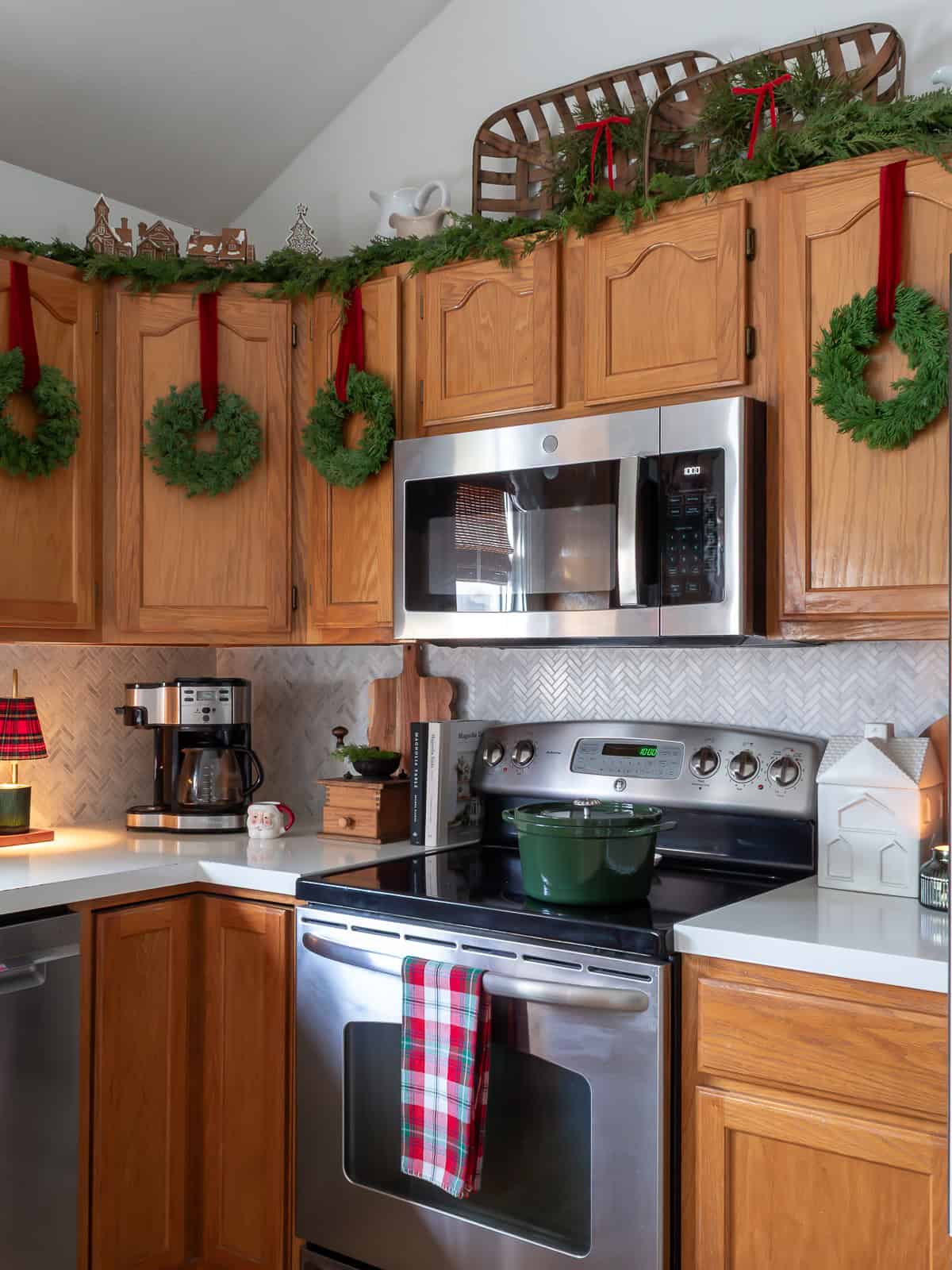 Kitchen decorated with traditional Christmas decorating ideas featuring wreaths hung on cabinets with red velvet ribbon, garland and DIY gingerbread village houses above the cupboards, a plaid towel on the stove, a green Dutch oven, and cozy Ralph Lauren Christmas inspired touches throughout the space.