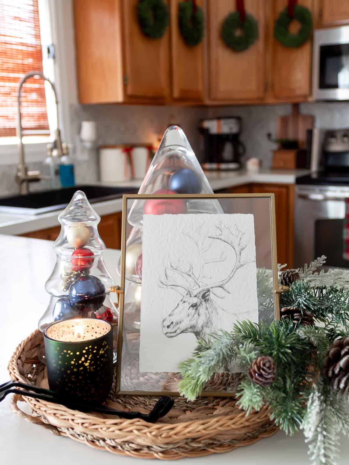 “Kitchen island vignette styled with traditional Christmas decorating ideas, including glass tree jars filled with ornaments, a framed reindeer sketch, a lit green candle, and greenery with pinecones on a woven tray, with wreaths on the cabinets and Ralph Lauren Christmas inspired touches in the background.”