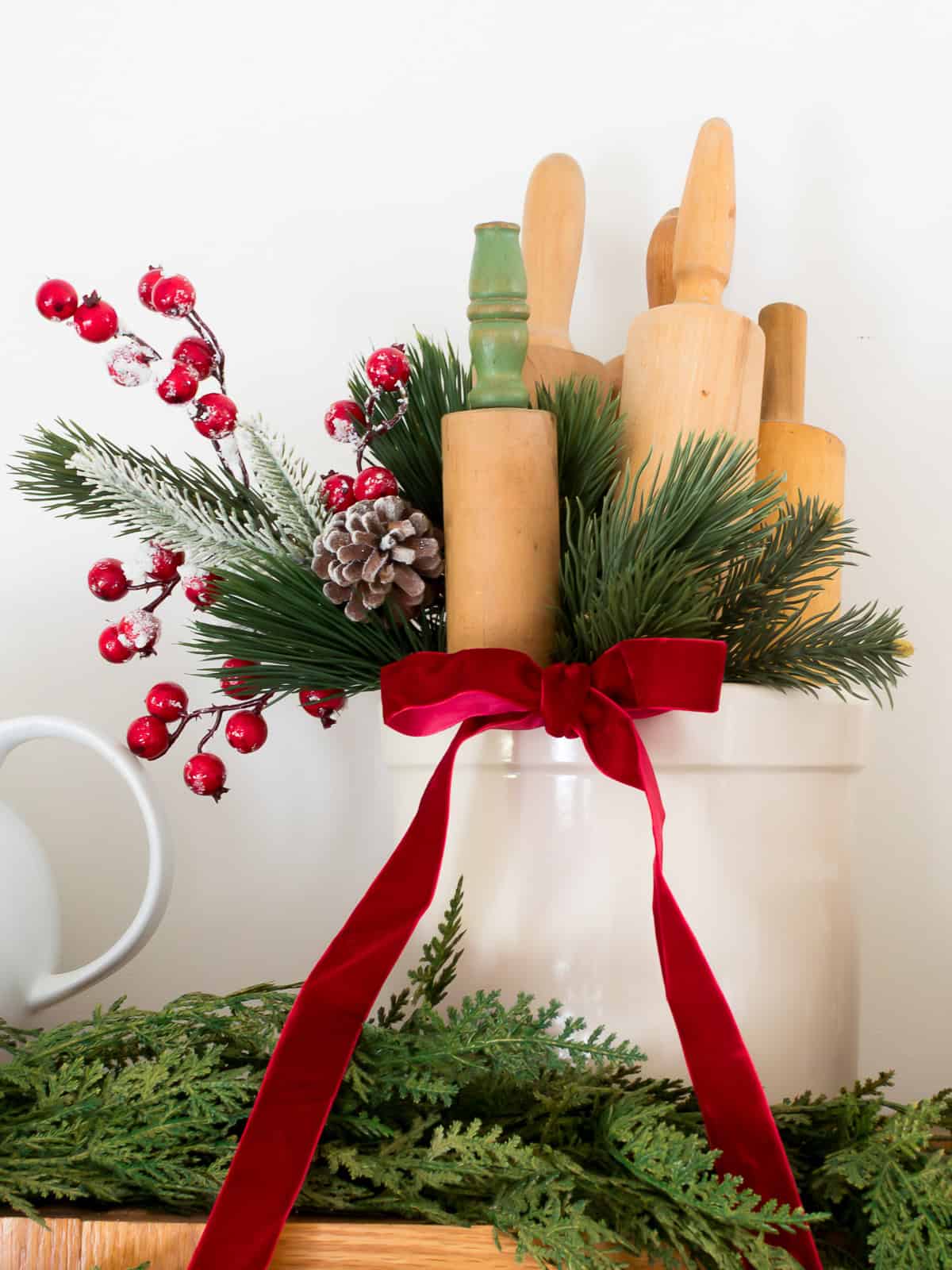 A vintage crock placed on top of kitchen cabinets filled with greenery and, a spring of red berries and pinecones, filled with vintage rolling pins and adorned with a red velvet bow