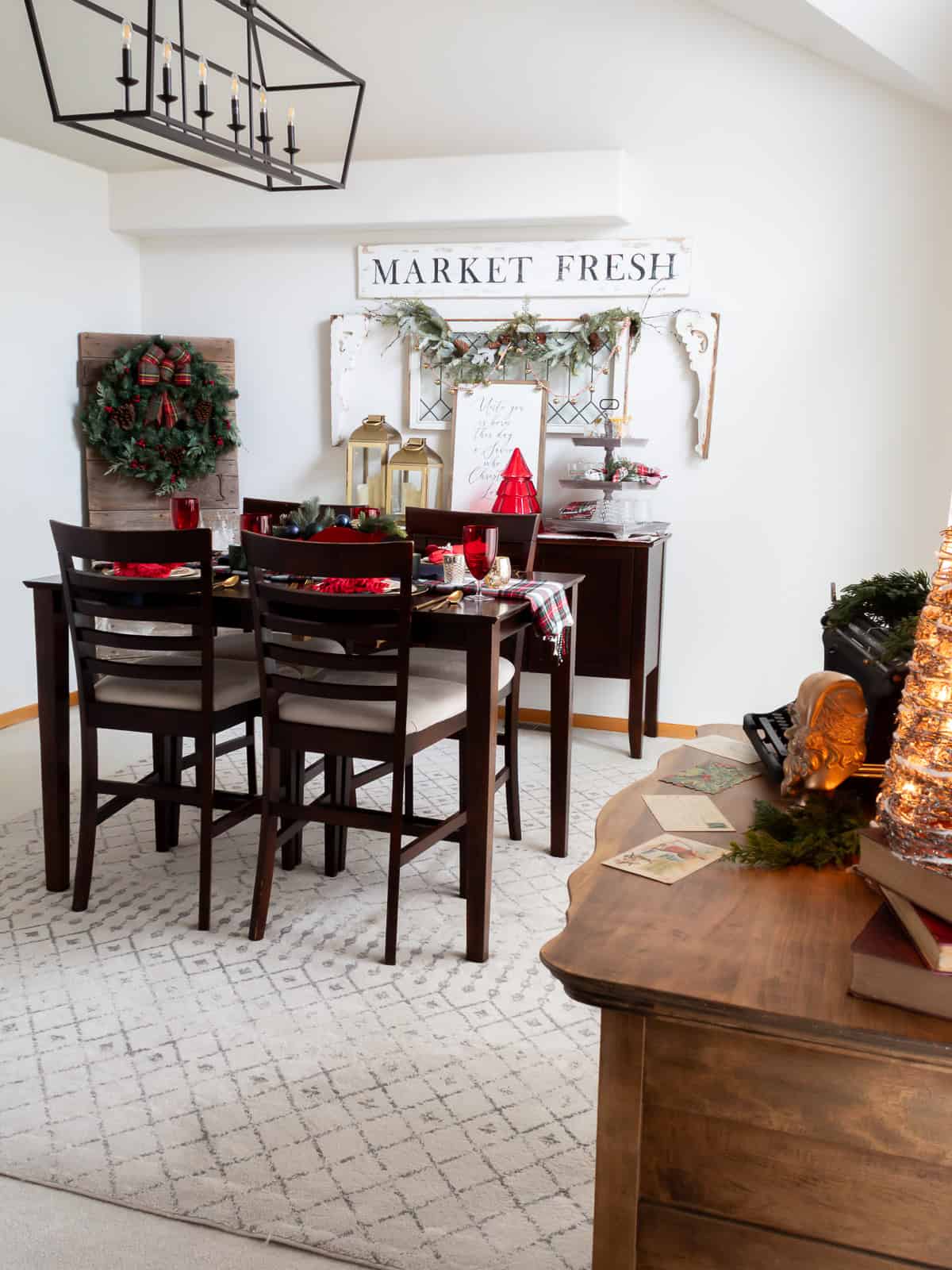 Dining room decorated with traditional Christmas decorating ideas, featuring a tartan tablescape, red glassware, a large wreath with plaid ribbon, and a buffet styled with greenery, lanterns, and vintage cottage accents. A “Market Fresh” sign and seasonal touches create a warm, classic Christmas look.