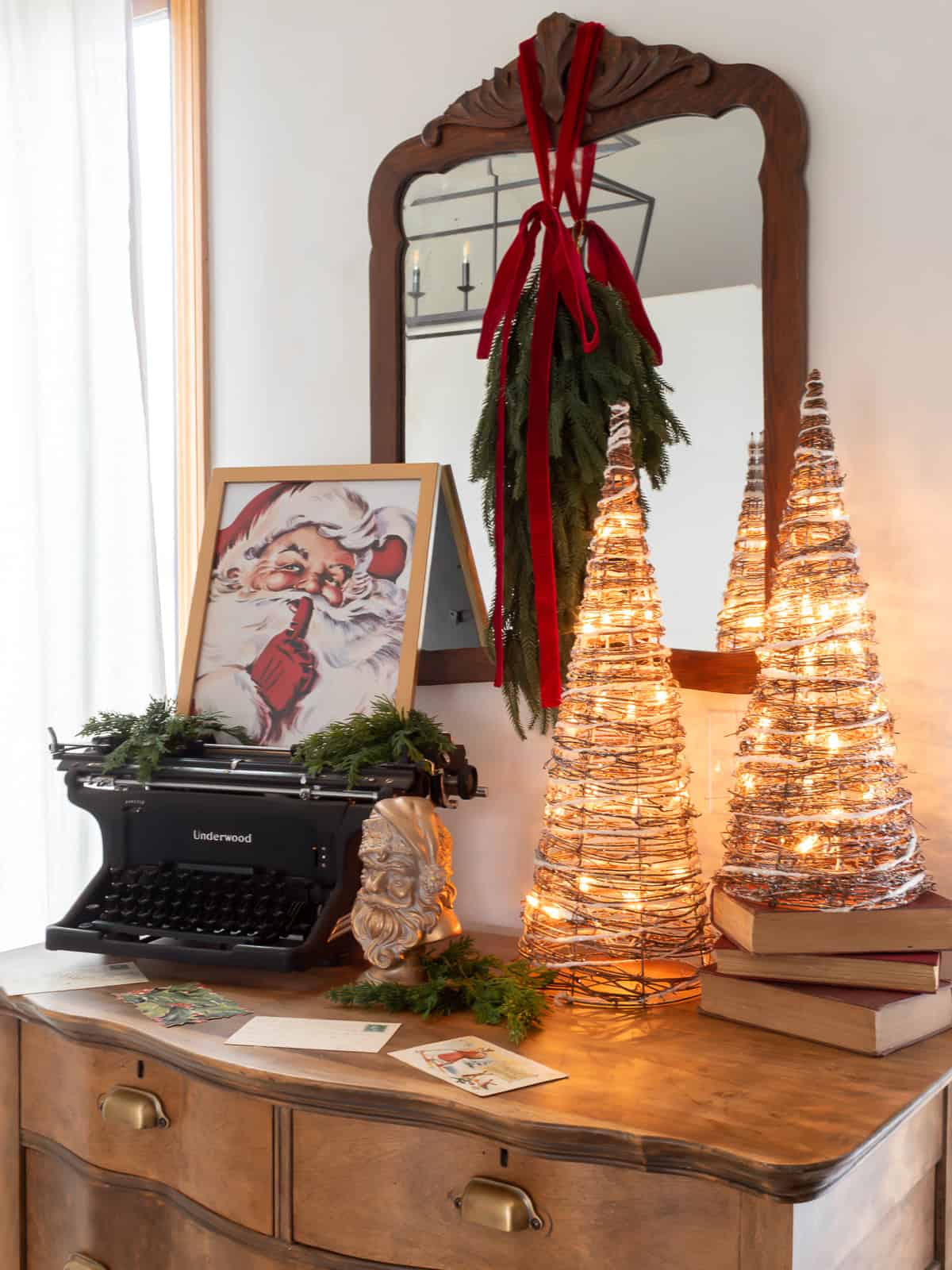 Traditional Ralph-Lauren inspired Christmas decorating ideas with a vintage dresser styled with an old typerwriter and framed santa print next to light up grapevine garaland cone trees and a vintage mirror on the wall with a christmas swag tied with red velvet ribbon