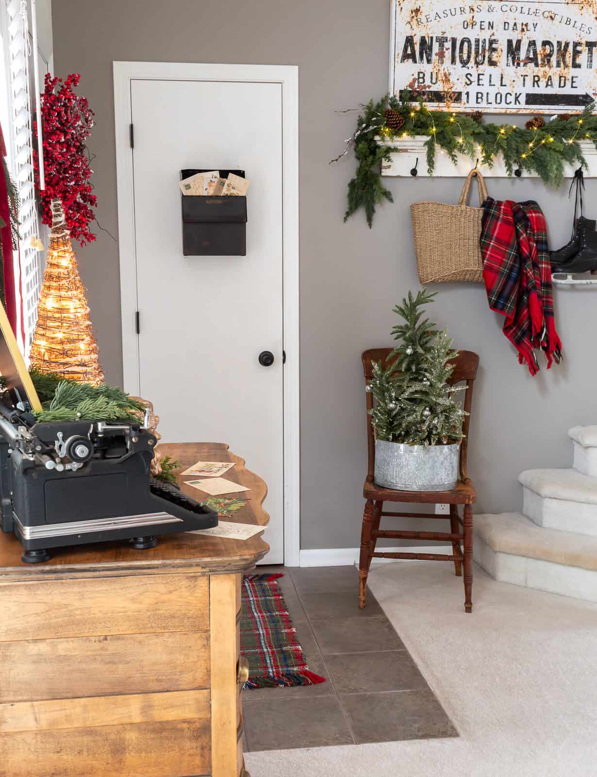 Entryway decorated with classic red and white Christmas decor, featuring a vintage dresser with a typewriter and postcards, a woven tree with lights, a red berry wreath, a black wall mailbox filled with vintage Christmas cards, a plaid throw, and cottage-style greenery
