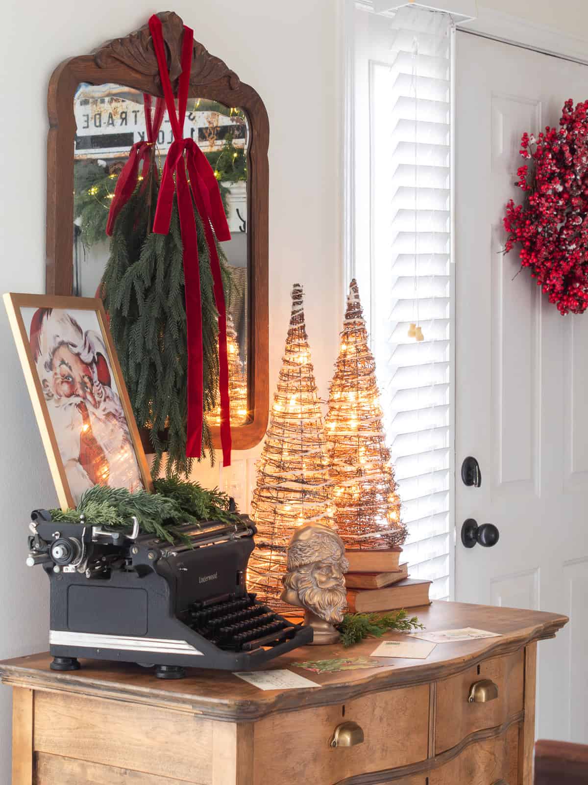 Close-up of a vintage dresser styled with traditional Christmas decorating ideas, including an antique typewriter with greenery, a framed Santa print, warm lighted cone trees, stacked vintage books, a gold Santa bust, and a mirror hung with red velvet ribbon, creating a cozy Ralph Lauren Christmas look