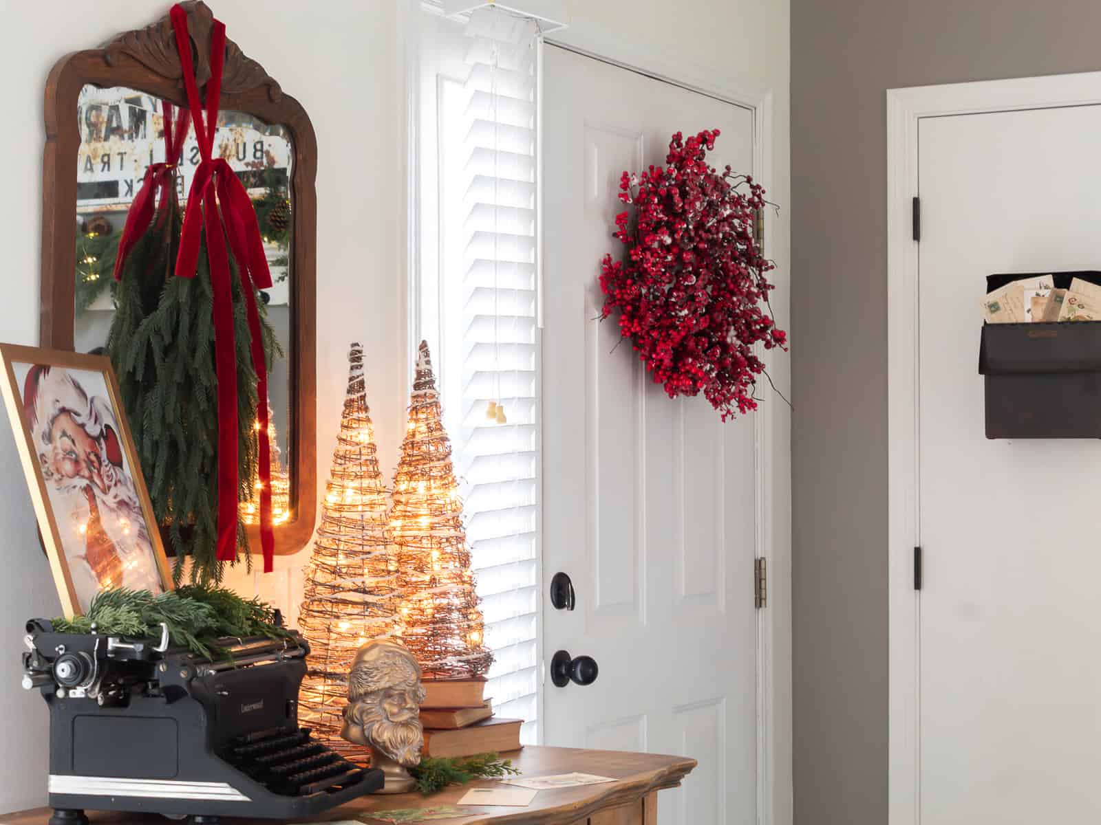 Entryway decorated with traditional Christmas touches, including a red berry wreath on the door, glowing grapevine Christmas trees on a vintage dresser, an antique typewriter with greenery, a framed Santa print, and a black wall mailbox filled with vintage holiday postcards.