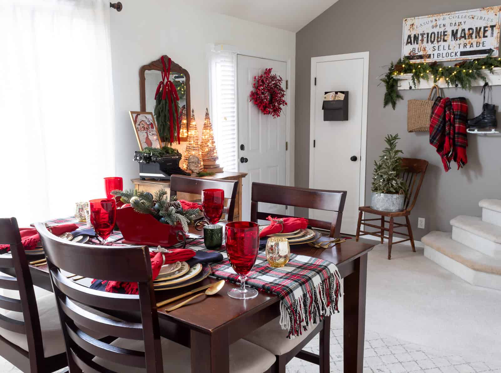 Dining room styled with traditional Christmas decorating ideas, including a tartan plaid table runner, red glassware, gold flatware, and a festive sleigh centerpiece. The entryway behind the table features classic Ralph Lauren Christmas accents like greenery, red ribbon, vintage decor, and warm, collected cottage-style touches.
