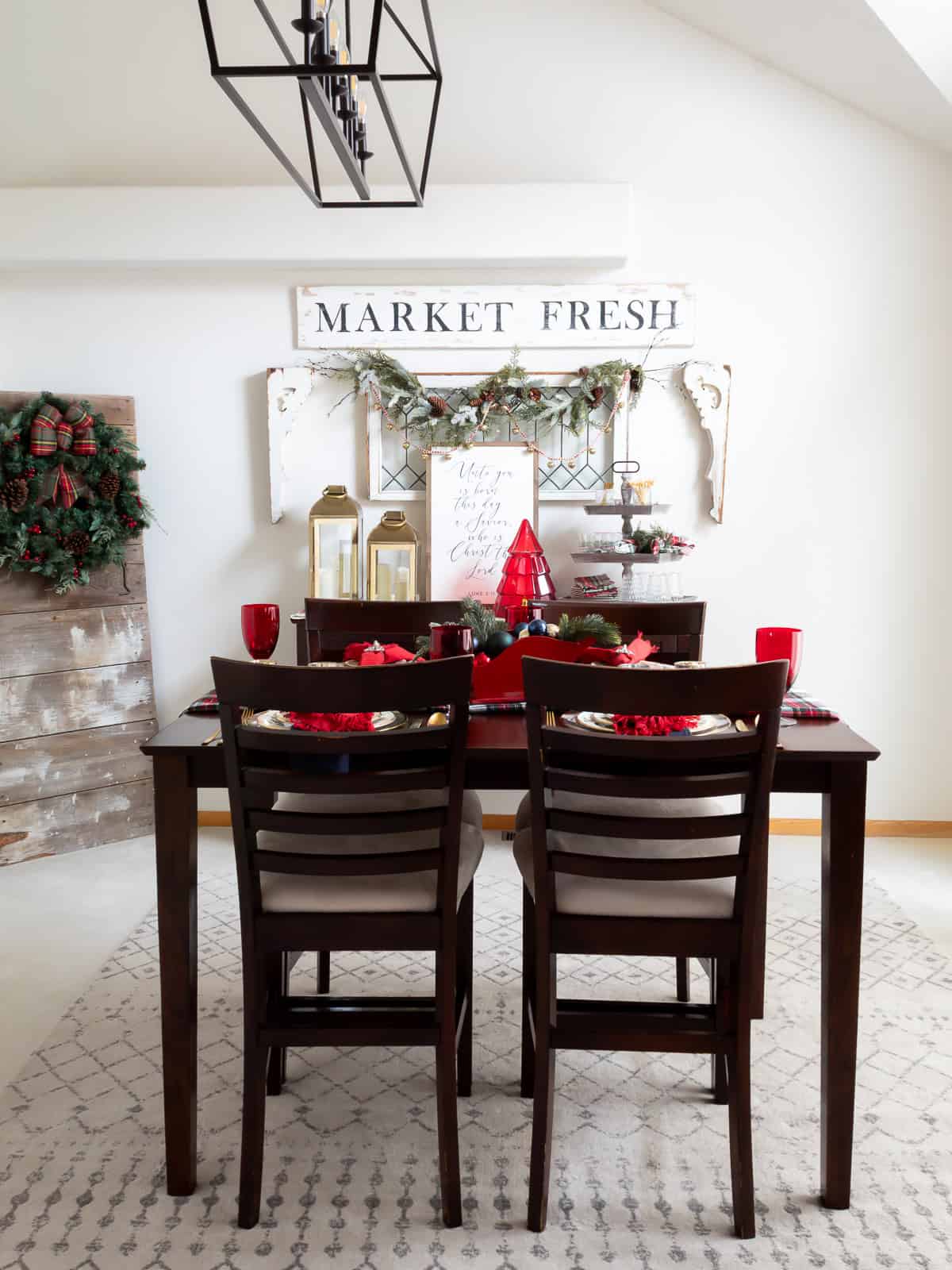 A festive dining room decorated for Christmas with a dark wood table set for two. The table is styled with red goblets, gold-rimmed chargers, tartan accents, and a sleigh centerpiece filled with greenery and ornaments. In the background, a buffet is decorated with gold lanterns, a red tree-shaped drink dispenser, and a Luke 2:11 sign. A garland with bells hangs over a vintage leaded-glass window, and a large wreath with a plaid bow hangs on a rustic barn door to the left.