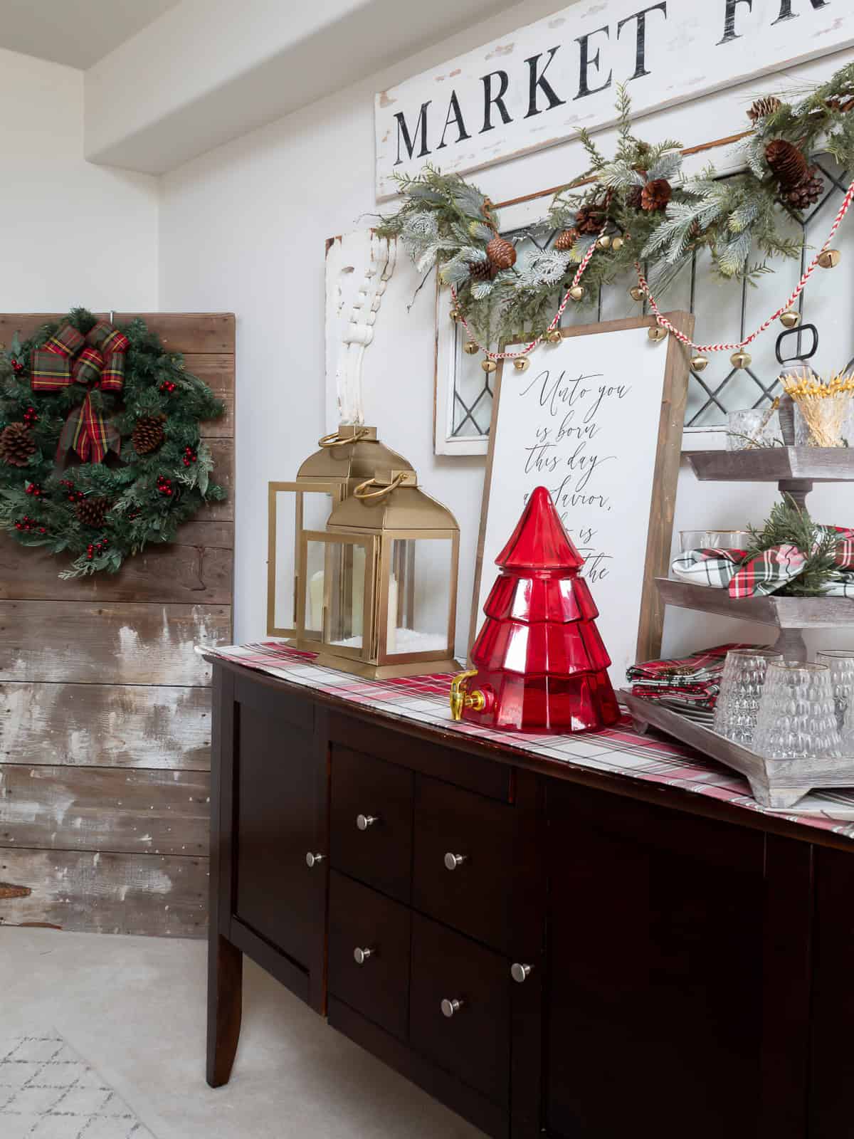 A festive dining room buffet decorated with traditional Christmas decorating ideas, including gold lanterns, a red tree-shaped drink dispenser, plaid linens, and a vintage leaded glass window topped with garland and jingle bells. A large wreath on a rustic barn door adds classic holiday charm to the space.