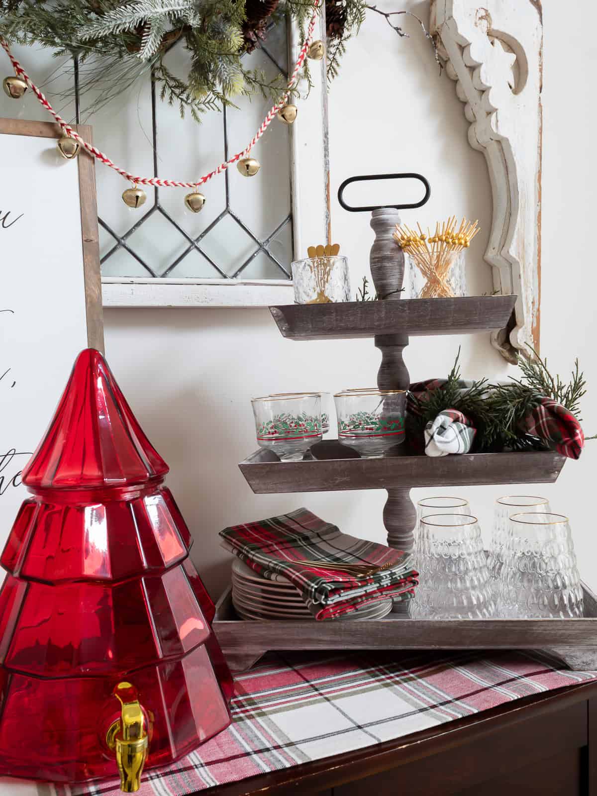 Tiered wooden tray on a dining room buffet styled with Christmas entertaining essentials, including vintage holiday glasses, plaid napkins, small plates, and greenery. A red Christmas tree drink dispenser sits beside the tray on a plaid runner, with garland and jingle bell decor hanging above.