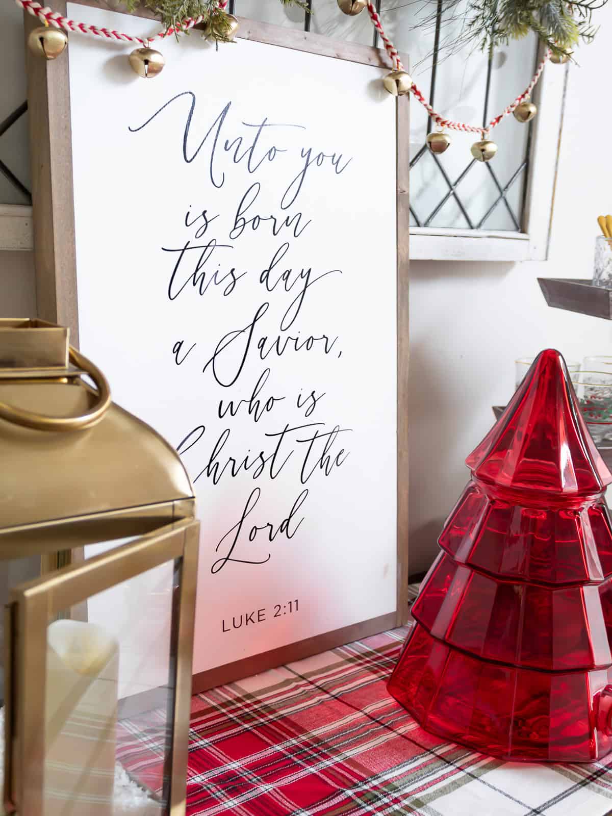 A Christmas buffet display featuring a Luke 2:11 Bible verse sign, a gold lantern with a candle, and a red glass Christmas tree drink dispenser, all styled on a red tartan plaid runner with greenery and jingle bell garland for a traditional holiday decorating look.