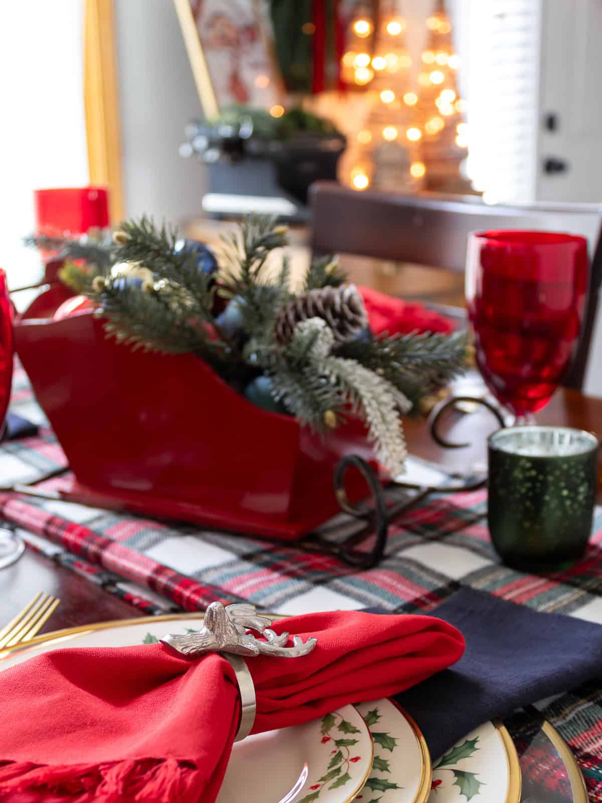 Festive dining table decorated for Christmas with a red sleigh centerpiece filled with greenery, pinecones, and ornaments, paired with red goblets and holiday dishes on a plaid blanket runner.
