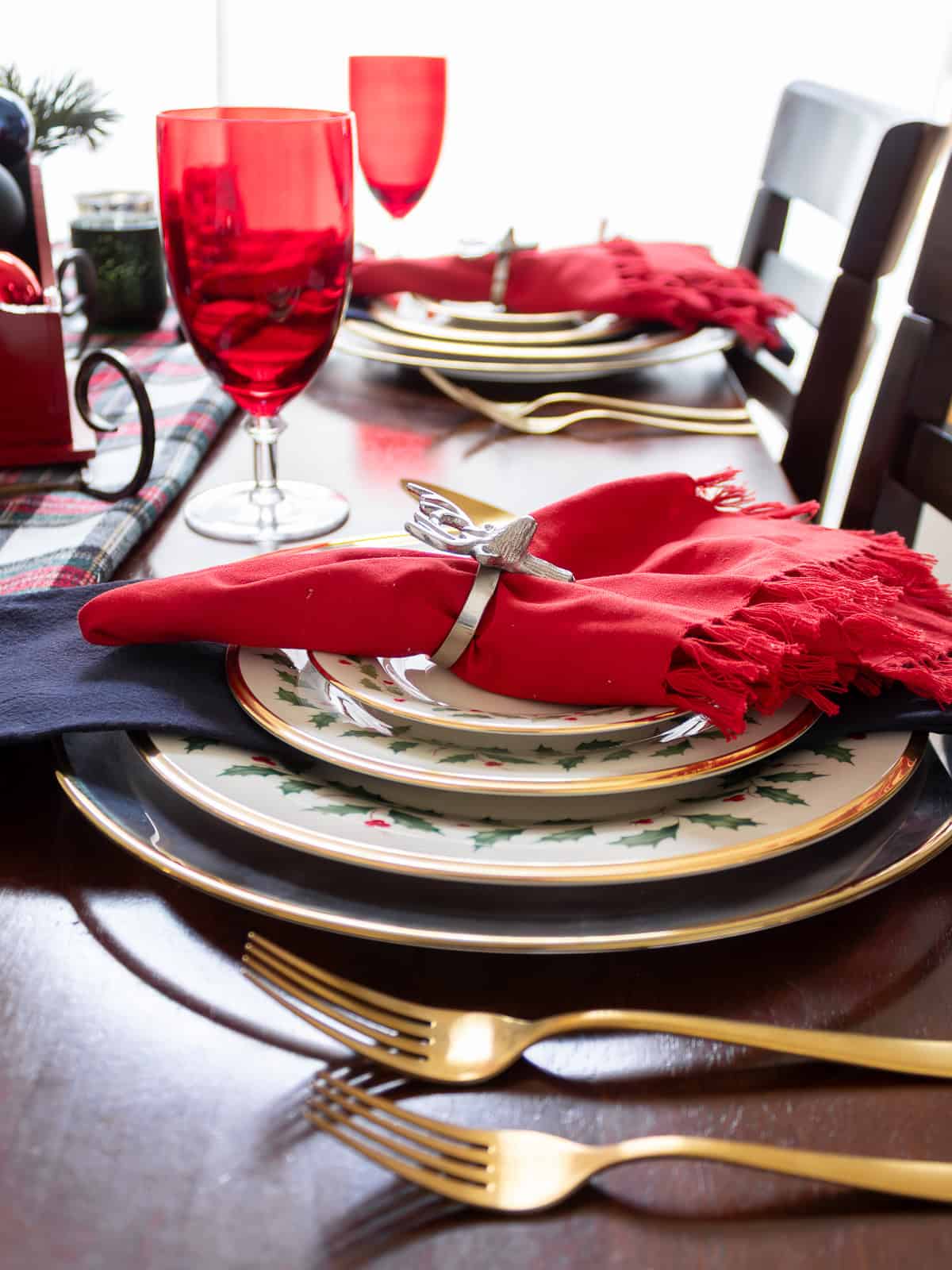 Close-up of a Christmas tablescape featuring red goblets, layered holiday plates, gold flatware, and a bright red fringed napkin with a silver deer napkin ring, all set on a plaid runner.