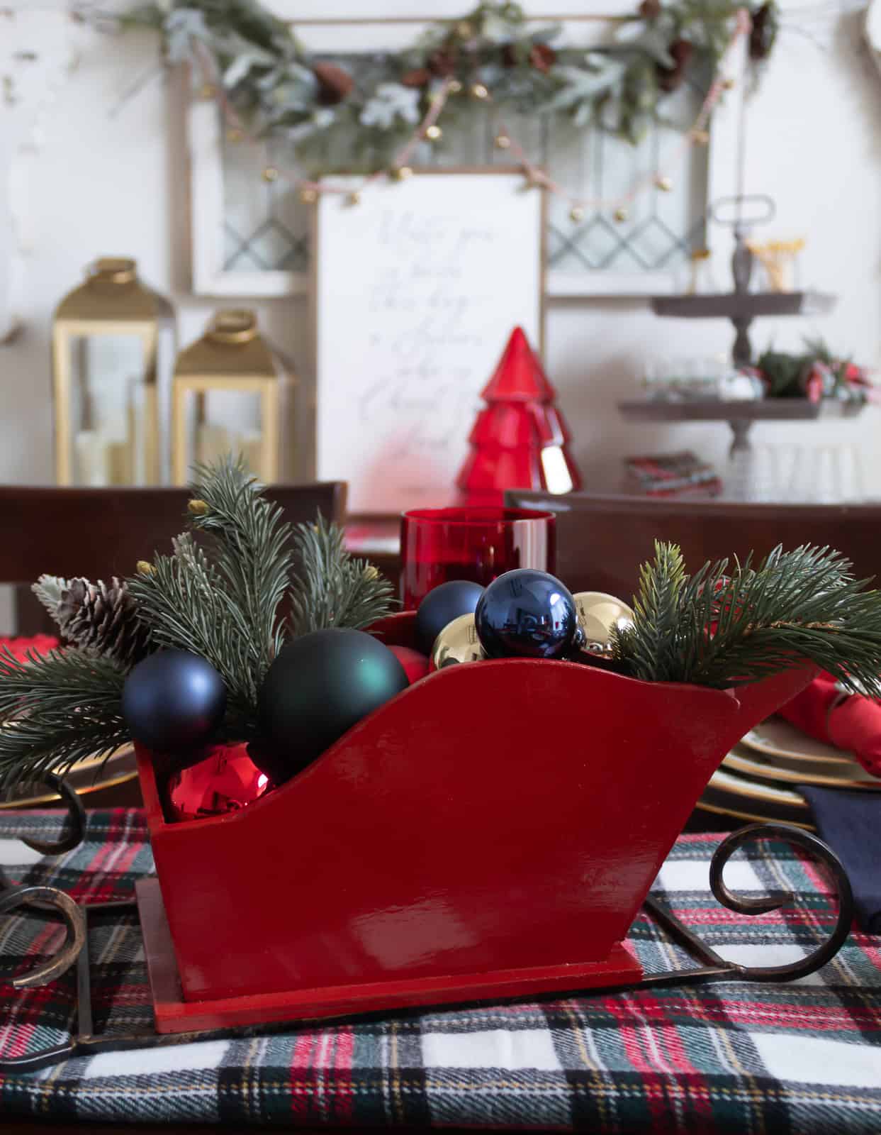 A festive red wooden sleigh centerpiece filled with evergreen sprigs, pinecones, and colorful ornaments sits on a plaid runner in the dining room. Red goblets, navy napkins, and soft holiday lights create a warm Christmas atmosphere, with the entryway decor and garland visible in the background.