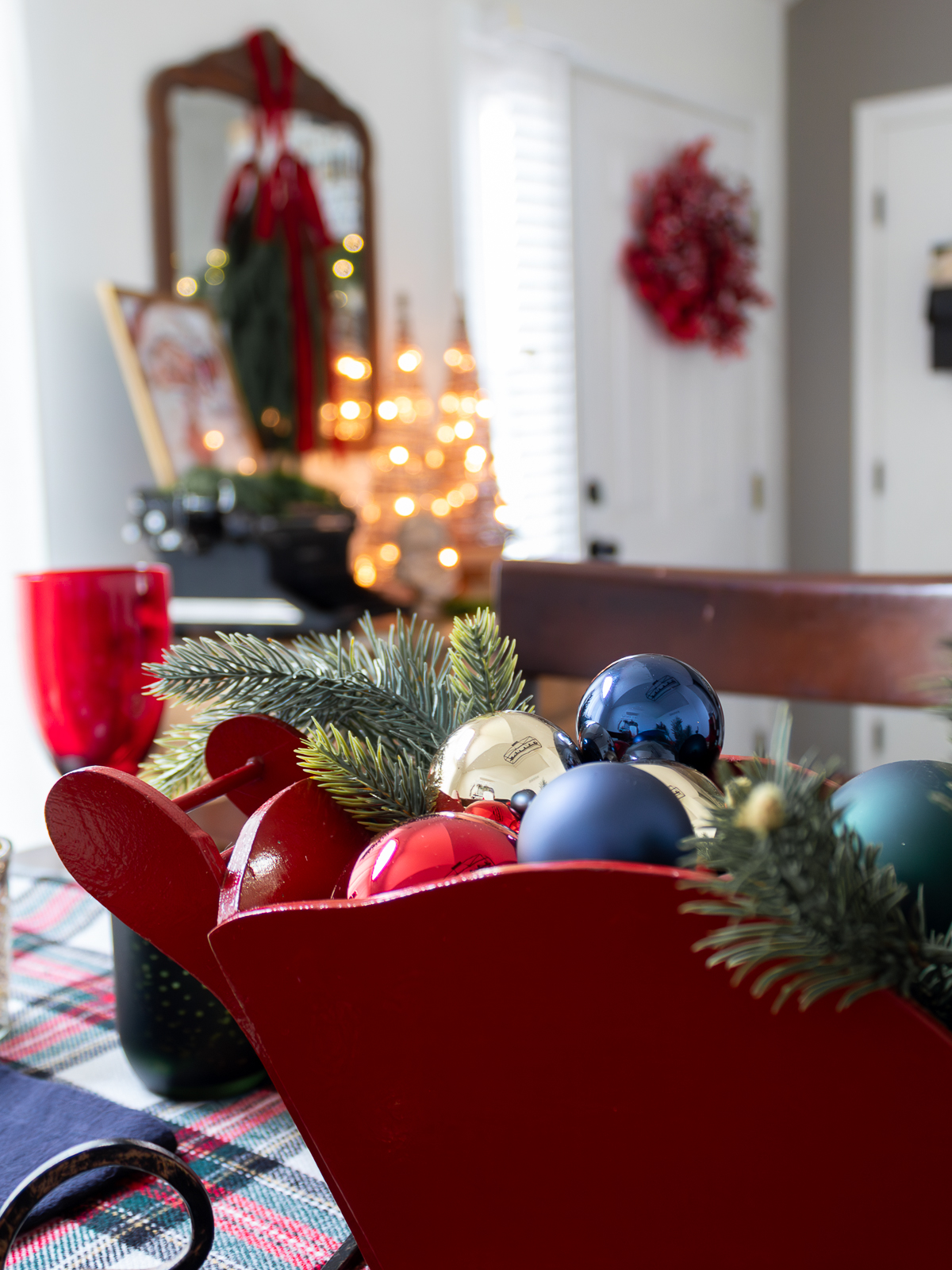 A close-up of a red wooden sleigh centerpiece on a Christmas dining table, filled with evergreen branches and red, gold, and navy ornaments. The plaid table runner and red glassware add a classic holiday touch, with softly glowing Christmas lights and a vintage mirror blurred in the background.