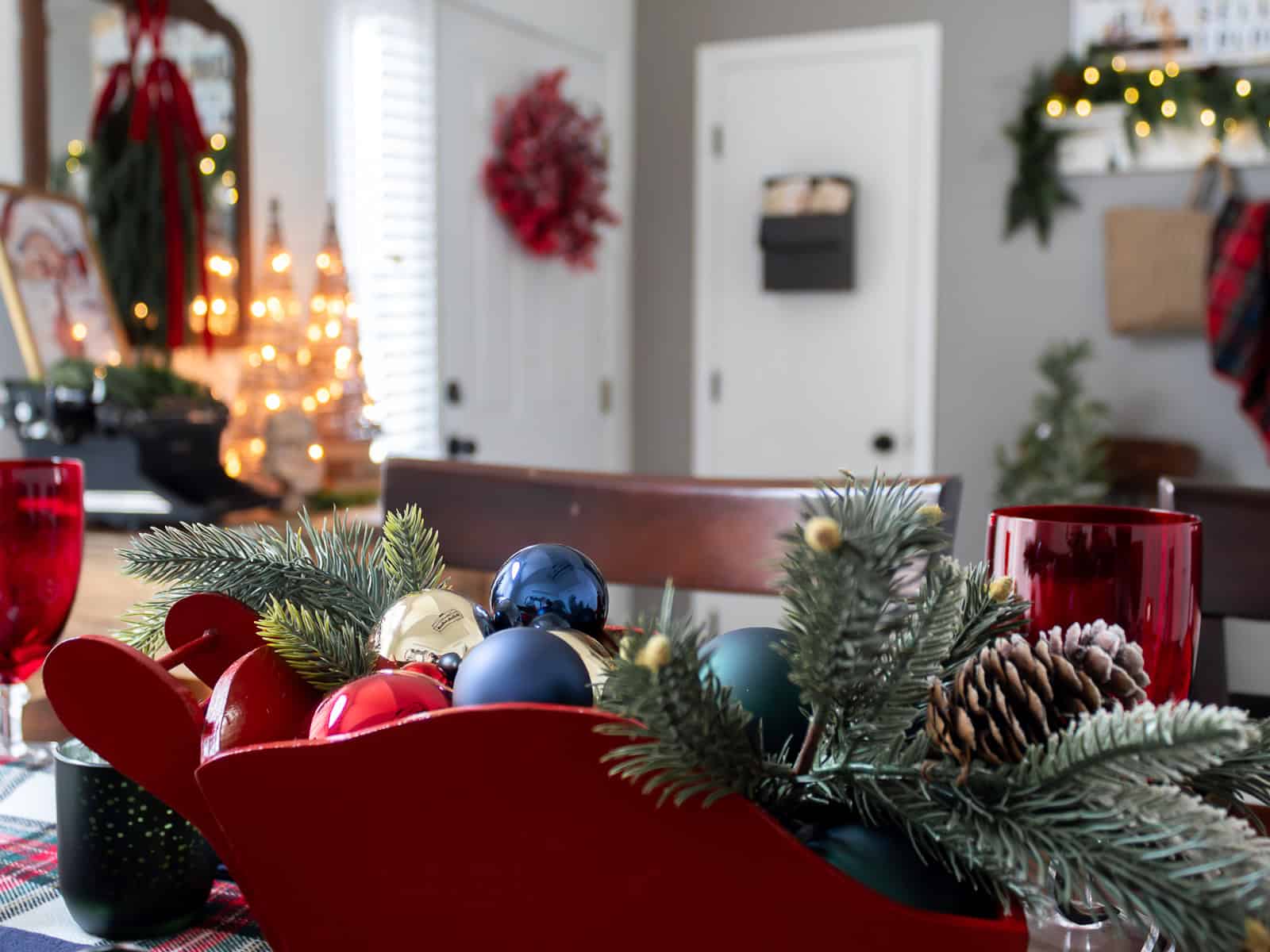 Close-up of a red wooden sleigh centerpiece on a Christmas tablescape, filled with evergreen branches, pinecones, and red, gold, and navy ornaments. The background shows a festive entryway with twinkle lights, a vintage mirror with a red bow, a berry wreath on the door, and holiday greenery along the wall hooks.