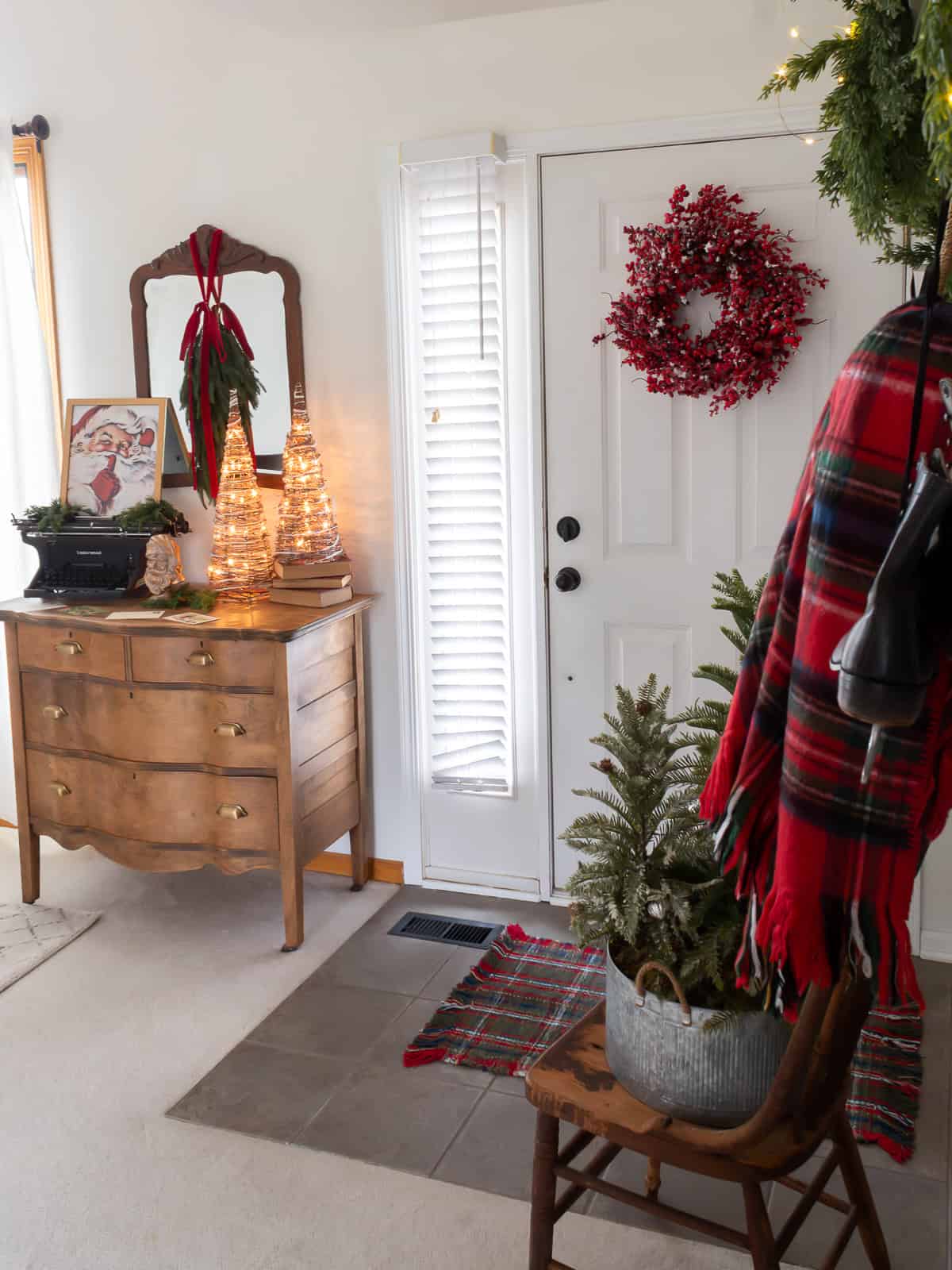 Entryway decorated with traditional Christmas decorating ideas featuring a vintage wooden dresser styled with a typewriter, Santa print, greenery, and lighted cone trees, a mirror hung with red velvet ribbon, a red berry wreath on the front door, and plaid accents for a cozy Ralph Lauren Christmas look