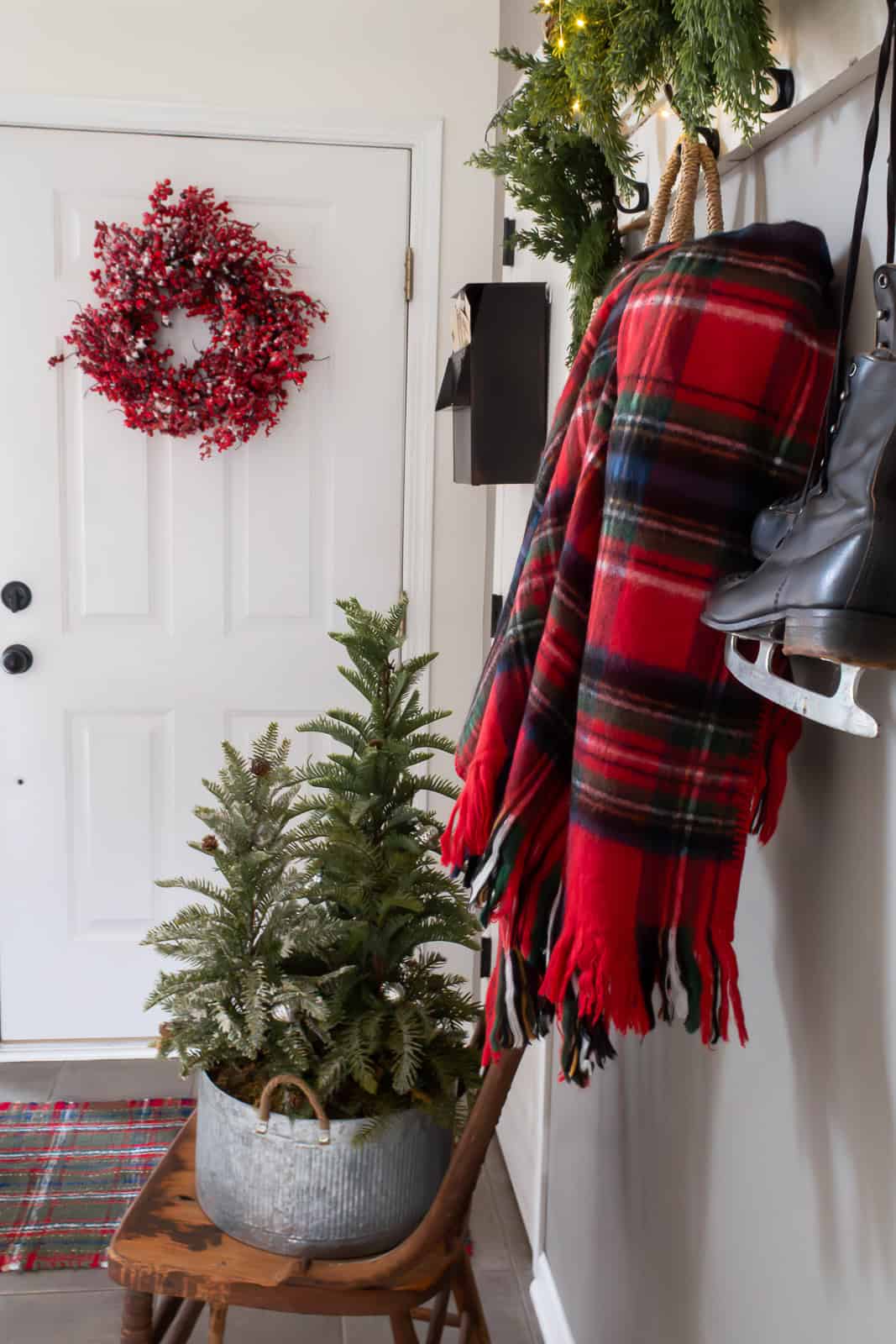 A welcoming Christmas entryway styled with traditional Christmas decorating ideas, including a vintage chair topped with a small festive tree, a tartan plaid throw, hanging ice skates, a mailbox filled with vintage postcards, and a red berry wreath on the door under a shelf decorated with greenery and warm twinkle lights.