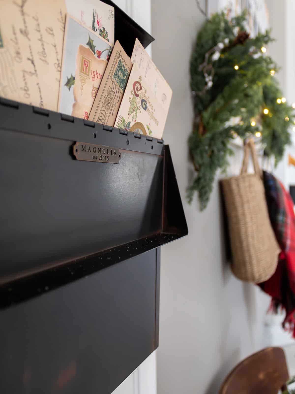 Close-up of a black wall-mounted mail organizer filled with vintage Christmas postcards, styled next to a holiday wreath with greenery and warm twinkle lights in a cozy entryway.