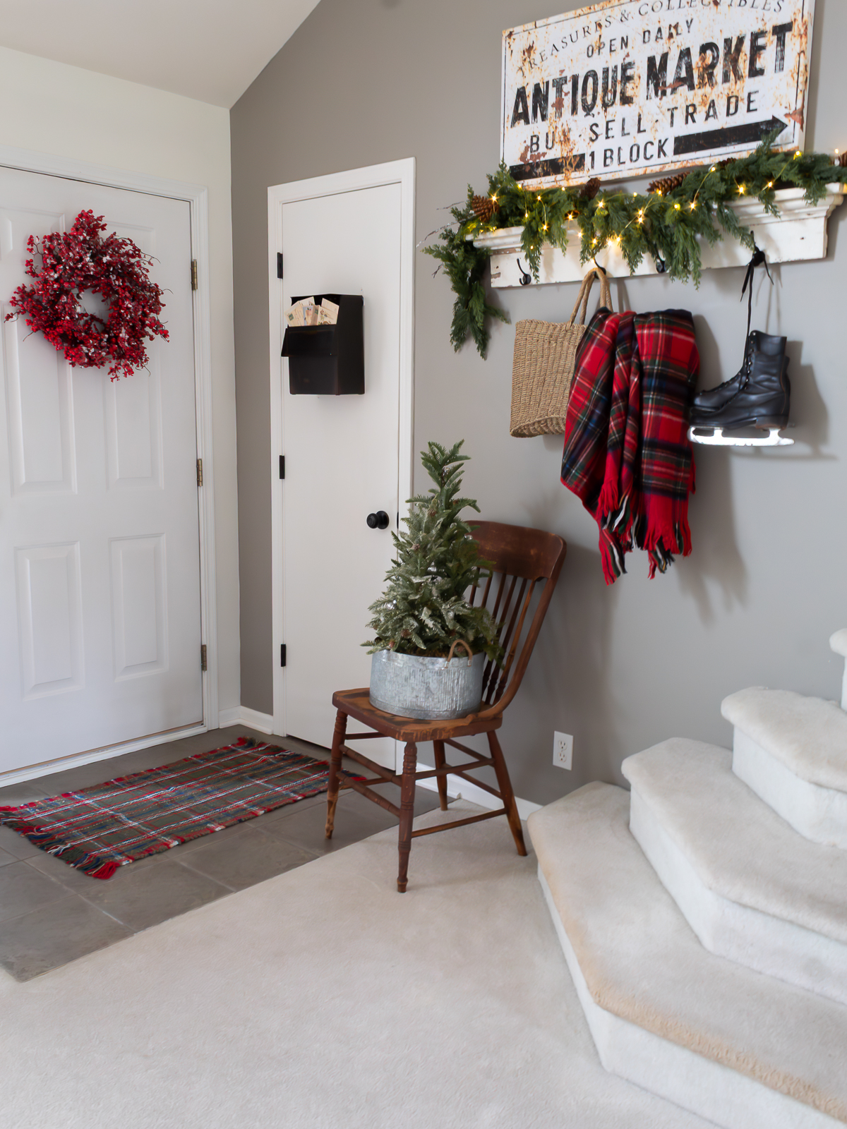 A cozy entryway decorated with traditional Christmas decorating ideas, featuring a red berry wreath on the front door, a vintage wooden chair holding a small frosted Christmas tree in a galvanized bucket, and a tartan plaid blanket hanging from wall hooks with ice skates, greenery, and twinkle lights above.