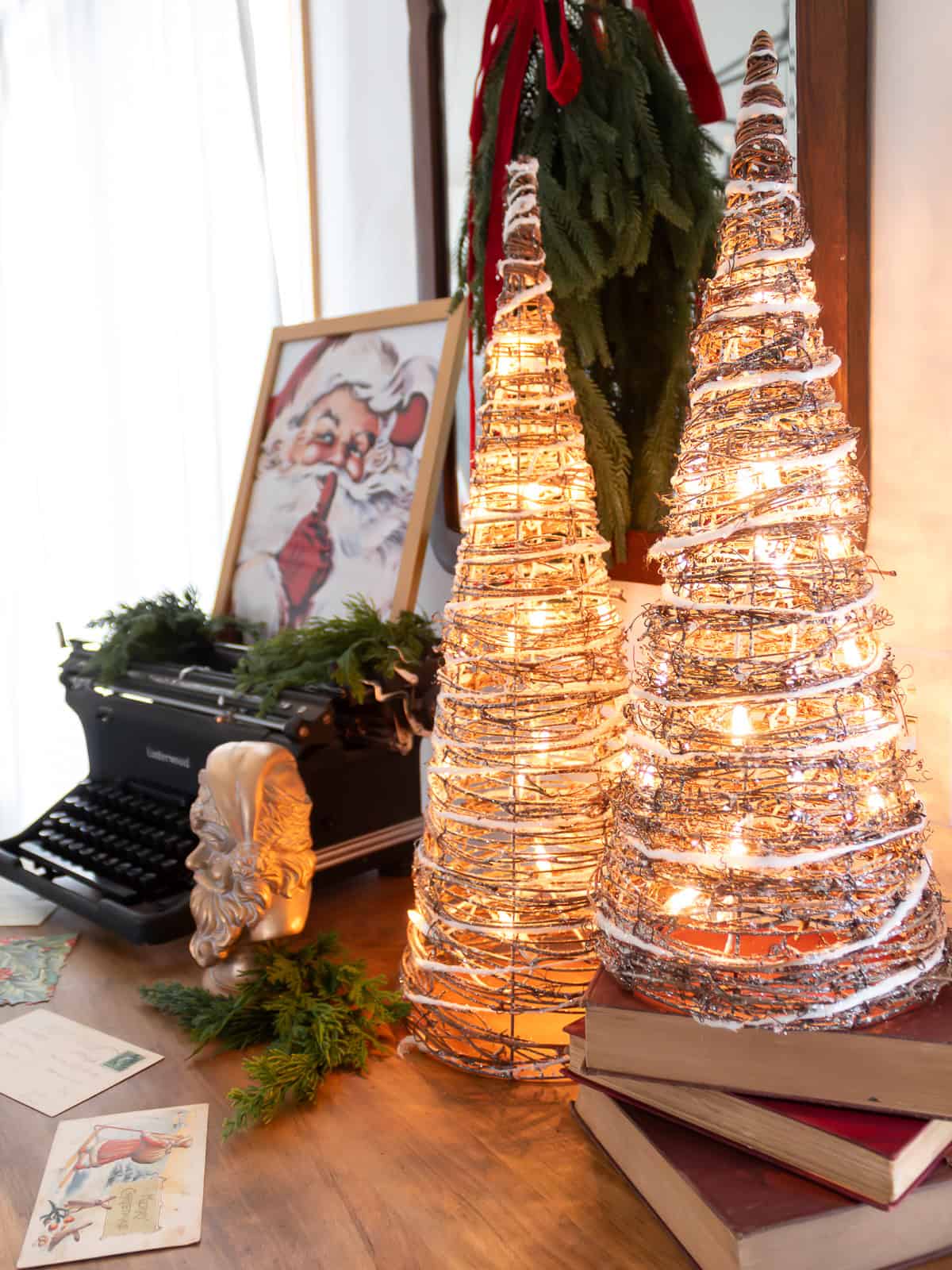 Close-up of a cozy Christmas vignette featuring glowing grapevine Christmas trees, vintage books, a golden Santa bust with greenery, an antique typewriter, and scattered holiday postcards, creating a warm and nostalgic Christmas display.