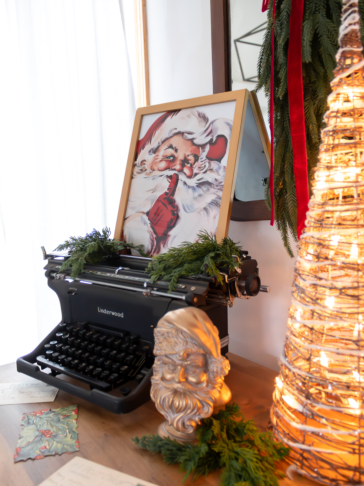 Traditional Ralph-Lauren inspired Christmas decorating ideas with a vintage dresser styled with an old typerwriter and framed santa print next to light up grapevine garland cone trees near a gold santa bust and a vintage mirror on the wall with a christmas swag tied with red velvet ribbon