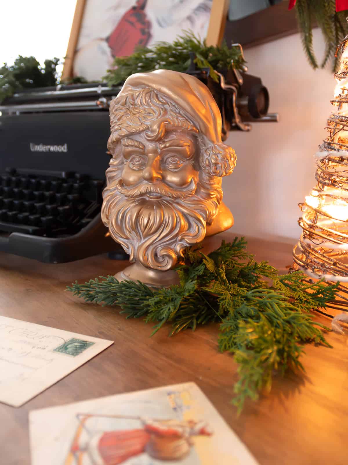 Close-up of a vintage gold Santa head figurine styled on a wooden entryway table with fresh greenery, antique Christmas postcards, warm twinkle lights, and a black Underwood typewriter, creating a cozy traditional Christmas decorating vignette.