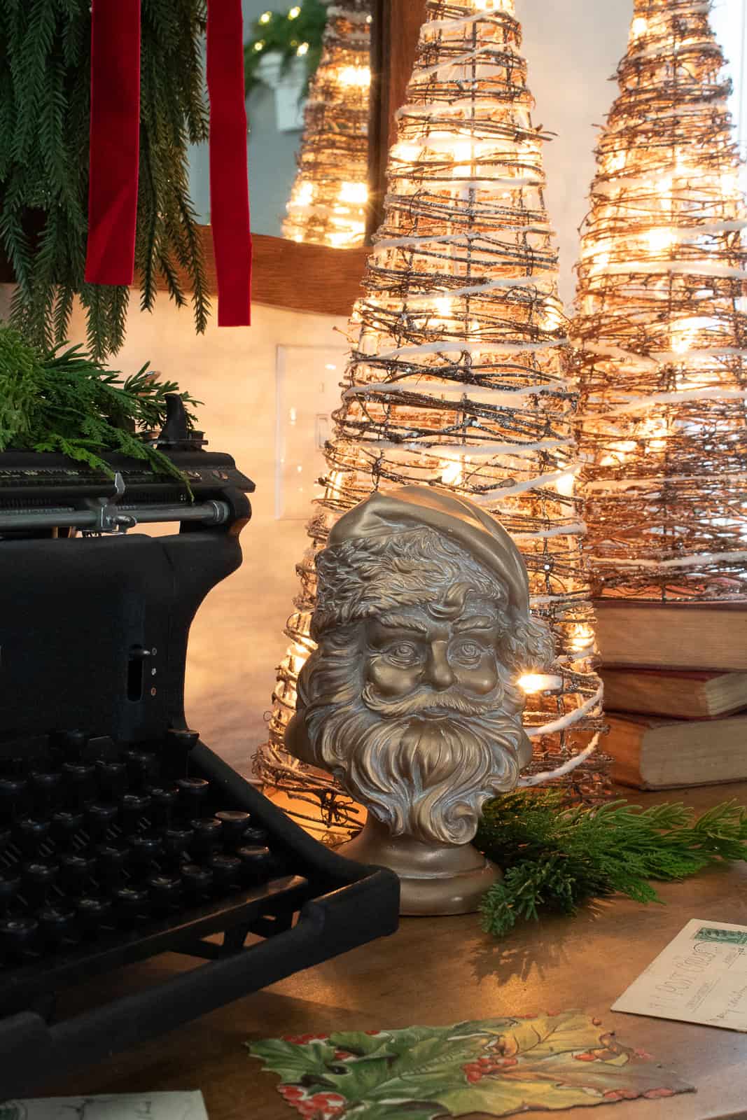 Close-up of a vintage gold Santa head figurine styled on a wooden entryway table with fresh greenery, antique Christmas postcards, warm twinkle lights, and a black Underwood typewriter, creating a cozy traditional Christmas decorating vignette.